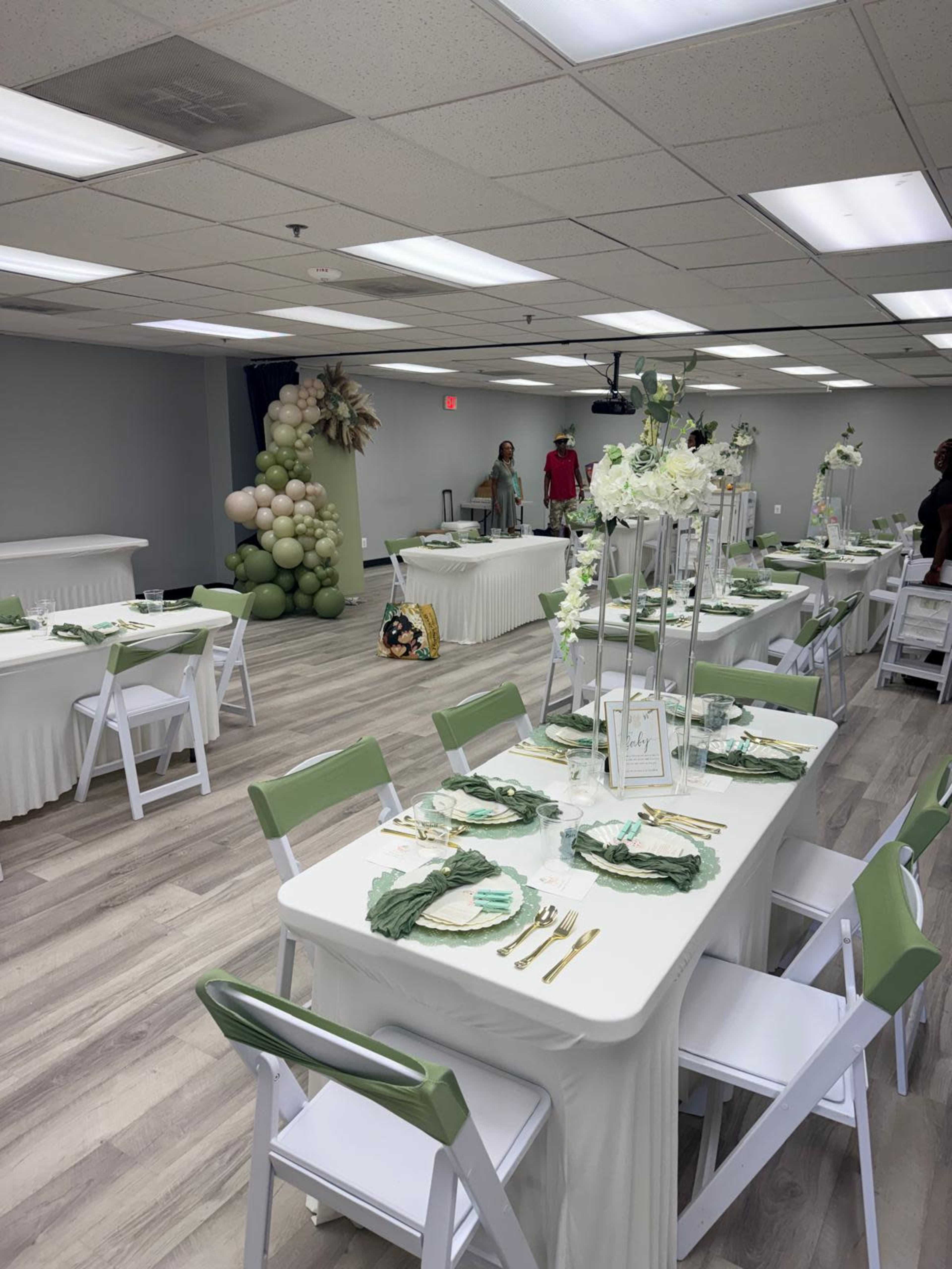 A well-decorated event space features multiple tables set with green and white tableware, complemented by floral arrangements and a balloon decoration in the background.