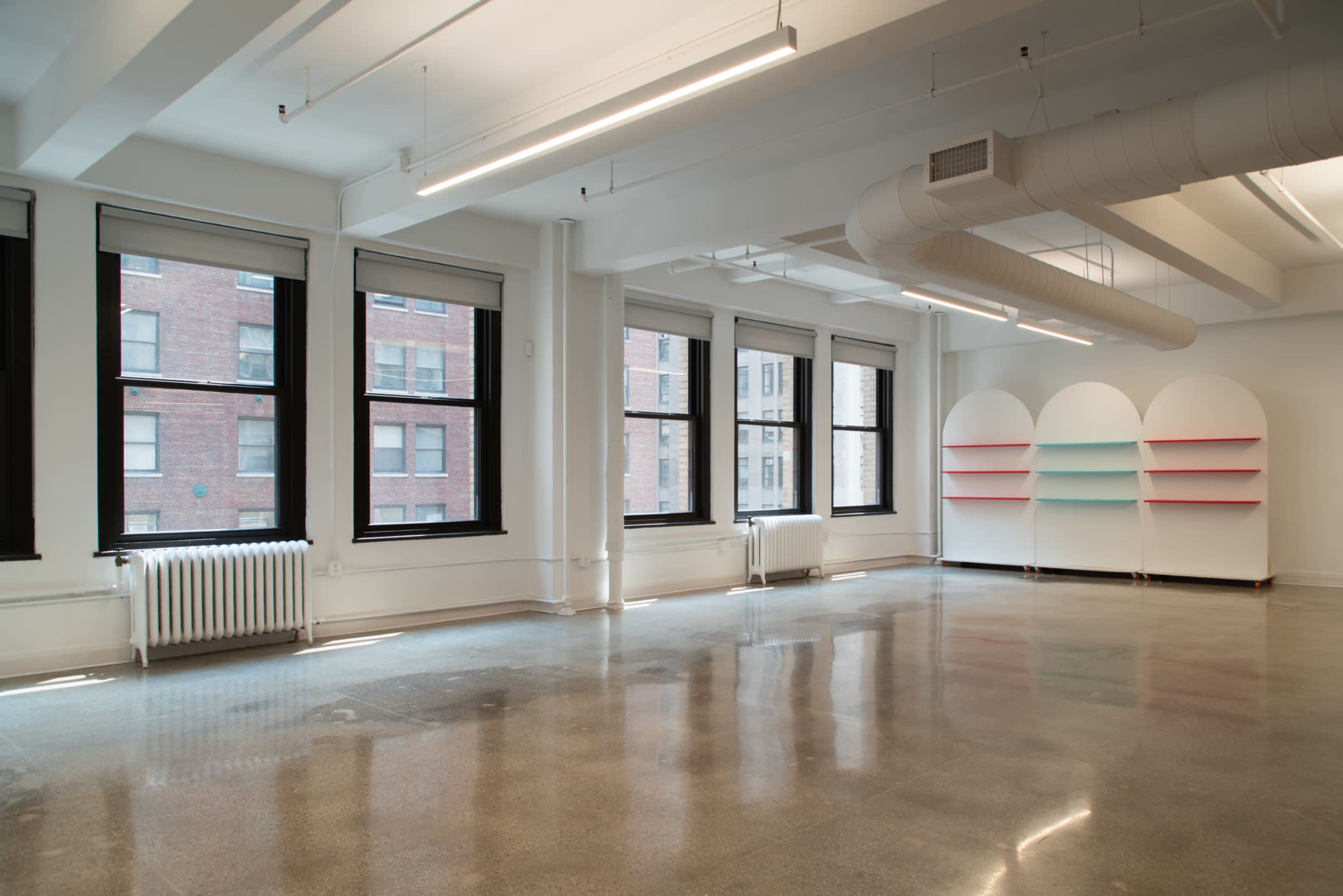 The image shows a spacious, empty room with large windows, concrete flooring, and a corner display unit with colored shelves.