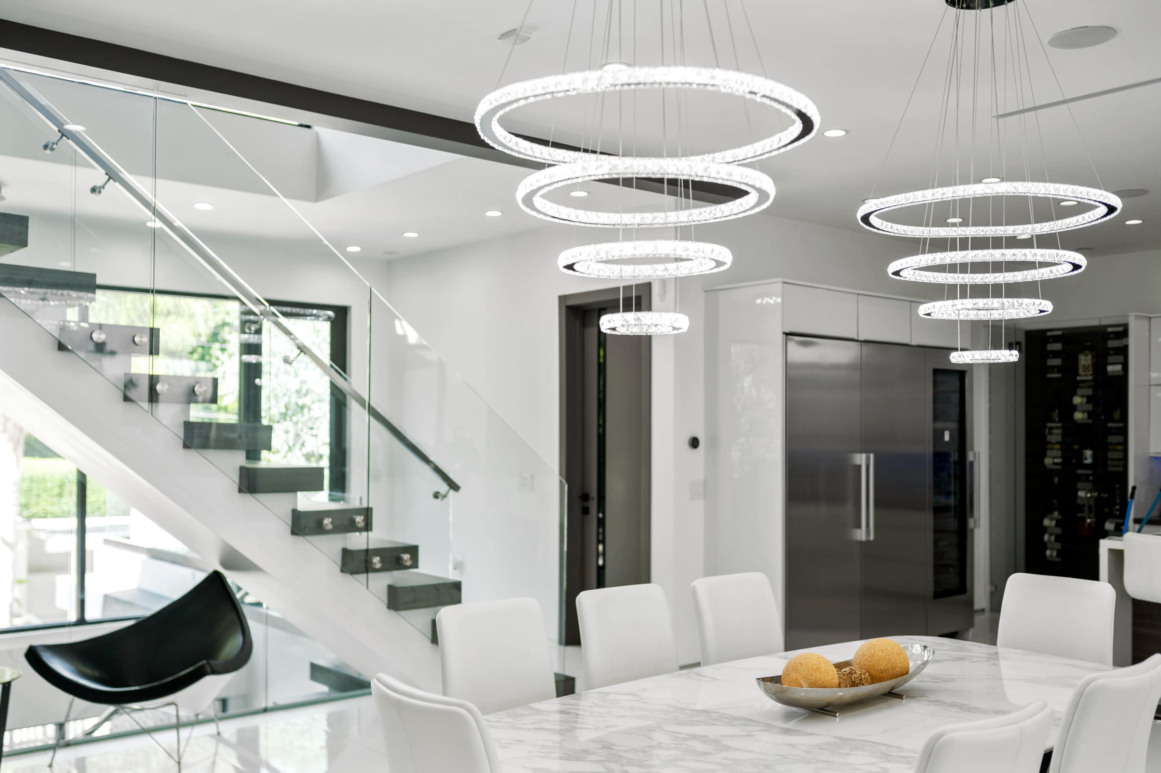 A modern dining area with a marble table, surrounded by white chairs, and illuminated by circular pendant lights, adjacent to a staircase and a glass wall.