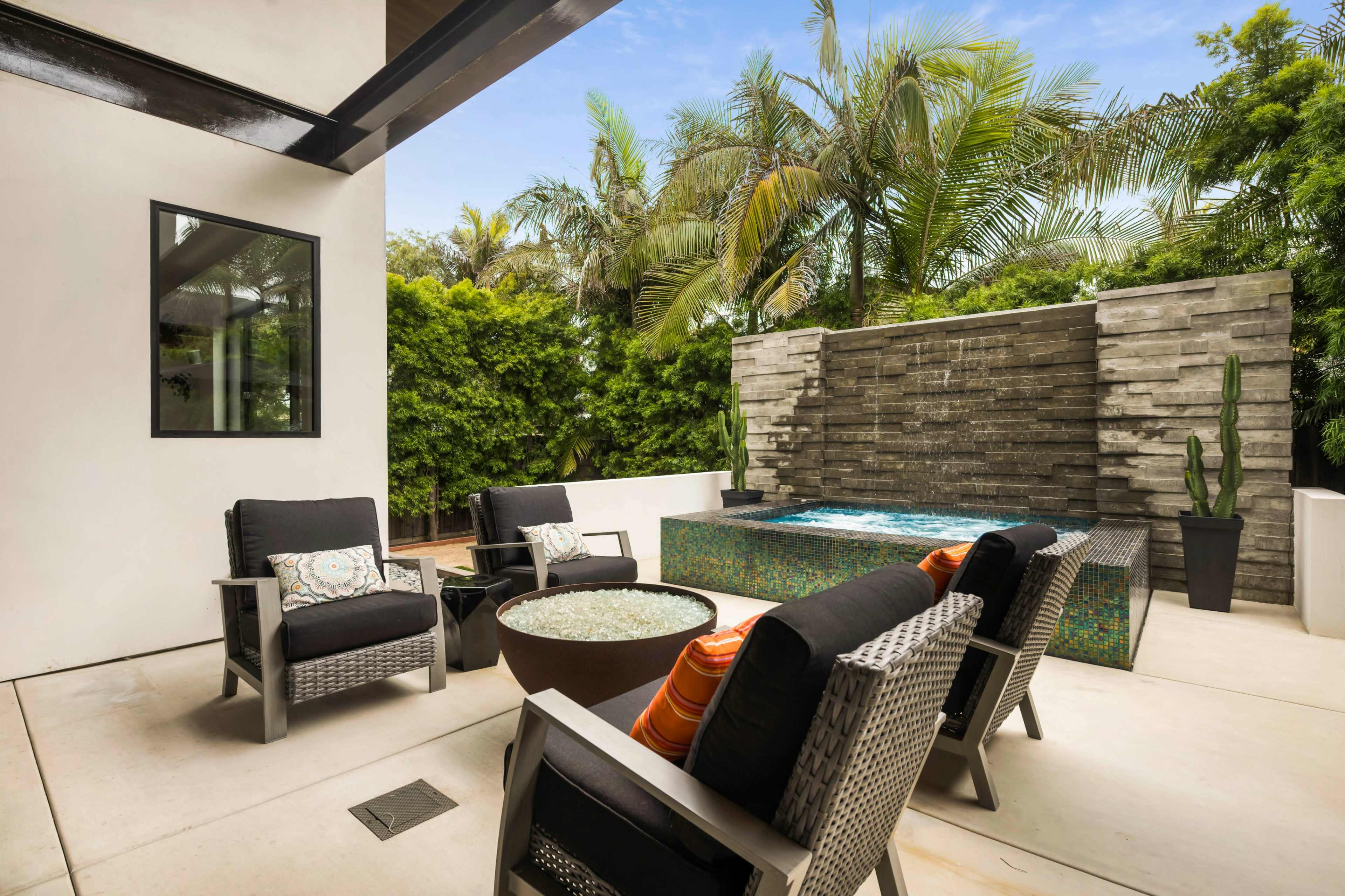 A modern outdoor patio features four lounge chairs around a fire pit, with a water wall and a small pond in the background surrounded by lush greenery.