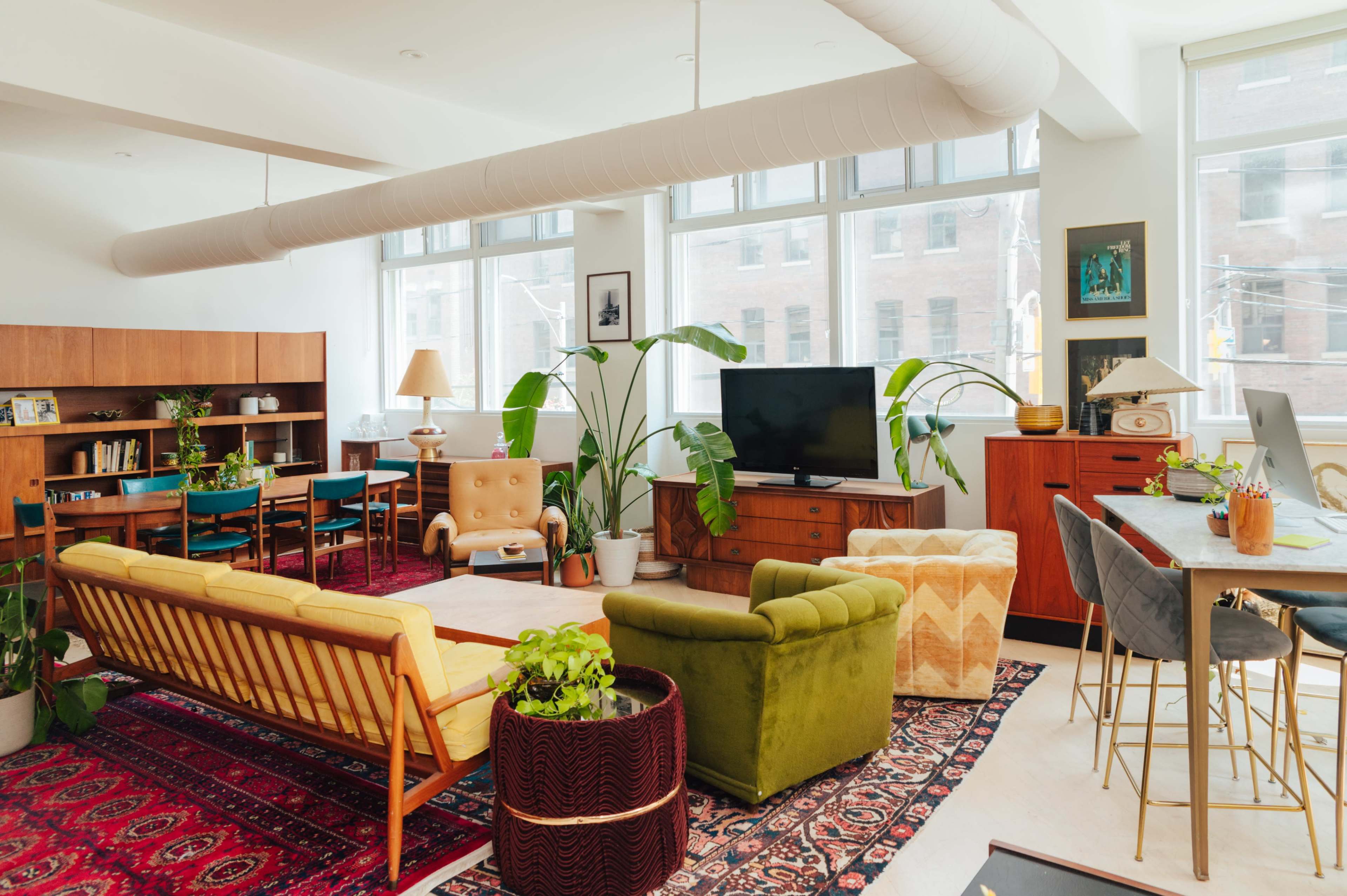 A modern living space featuring a mid-century style seating area, large windows, plants, and a variety of furniture including a television, bookshelves, and a dining area.