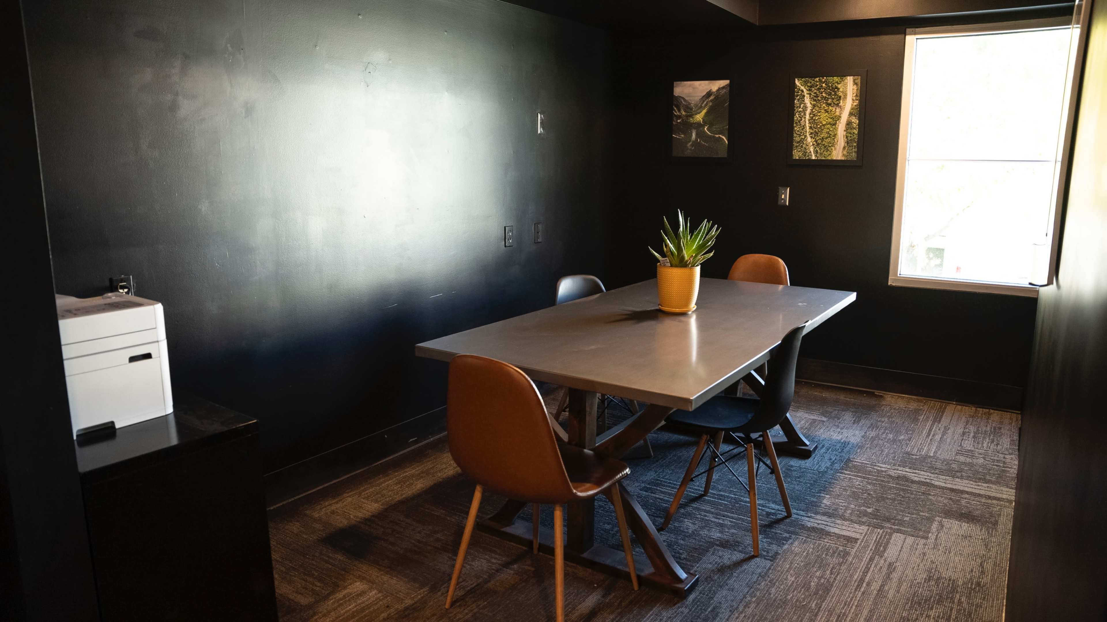 The image shows a small conference room with a table and four chairs, a potted plant in the center, and two framed pictures on the wall.