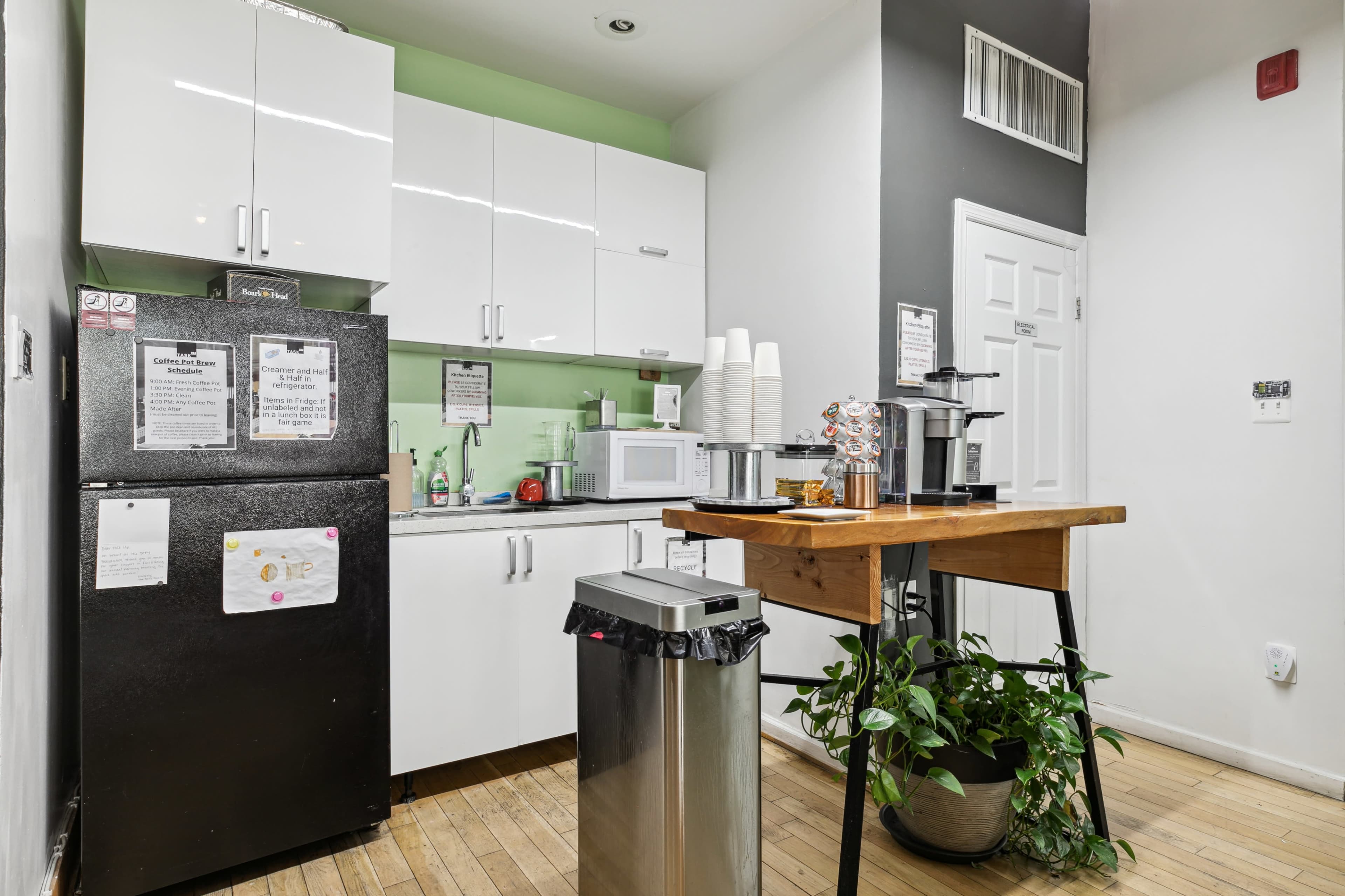 A modern kitchen area features a black refrigerator, white cabinets, a microwave, and a wooden table with a coffee machine, surrounded by a trash bin and a potted plant.