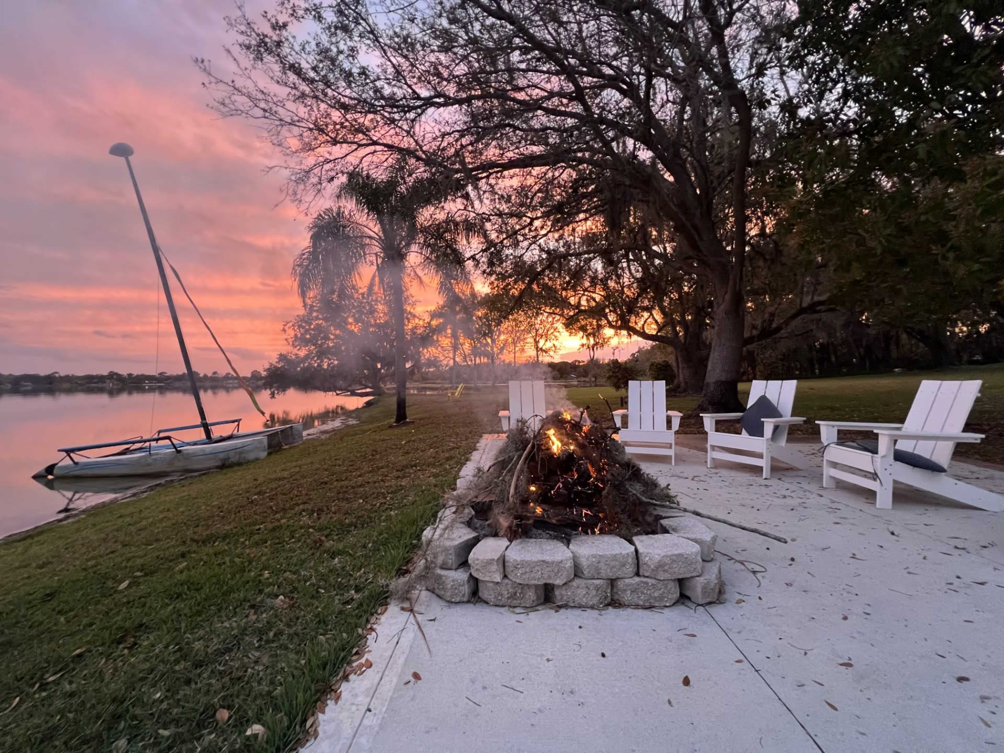 A fire burns in a stone circle beside a lake, with white outdoor chairs arranged around it and a sailboat moored nearby under a colorful sky at sunset.