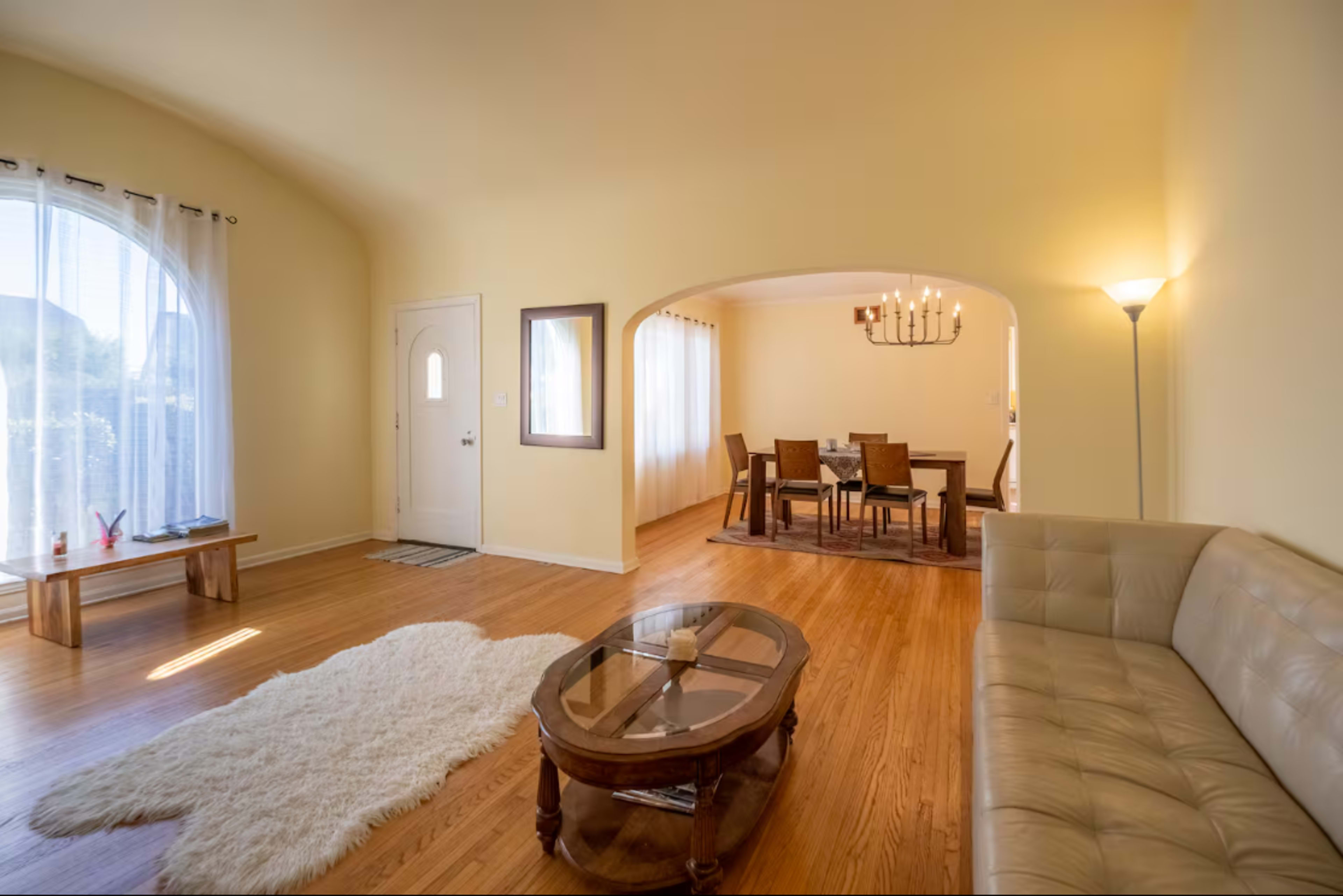 A cozy living room features a beige sofa, a wooden coffee table, a large area rug, and an archway leading to a dining area with a chandelier and wooden table.