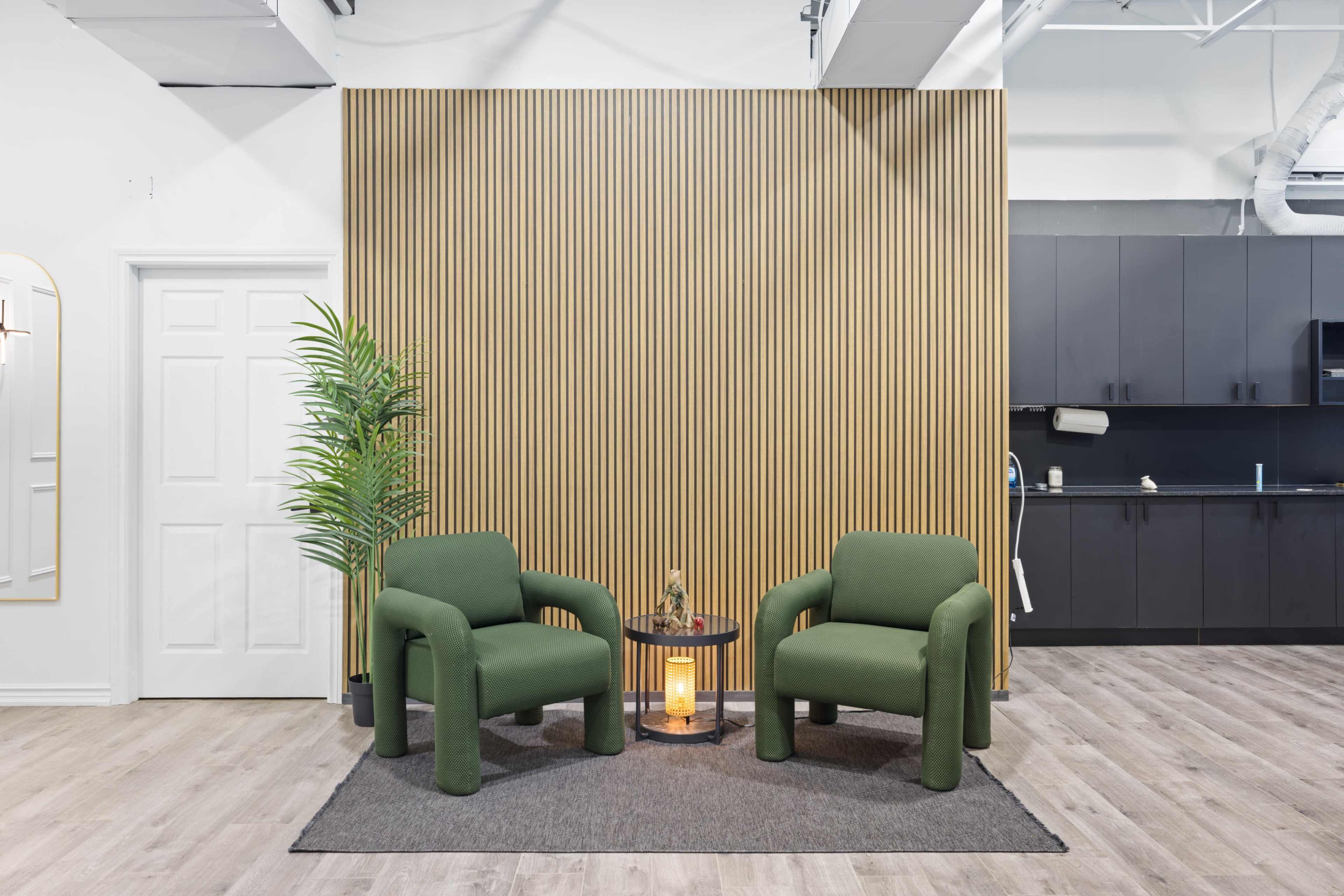 The image shows a modern interior with two green chairs facing each other, situated on a gray rug in front of a wooden slatted wall, with a small table and a kitchen area in the background.