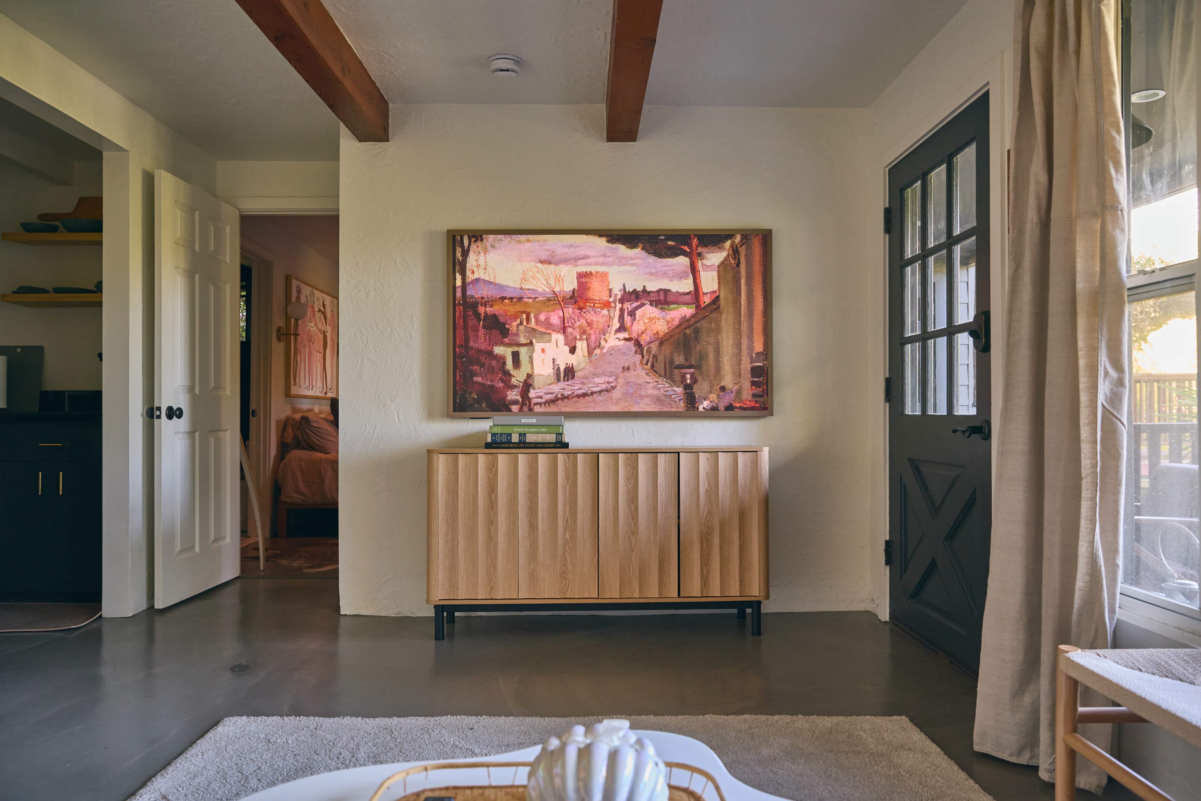 A wooden sideboard with a colorful painting above it sits in a modern living room, flanked by two doorways leading to other spaces.