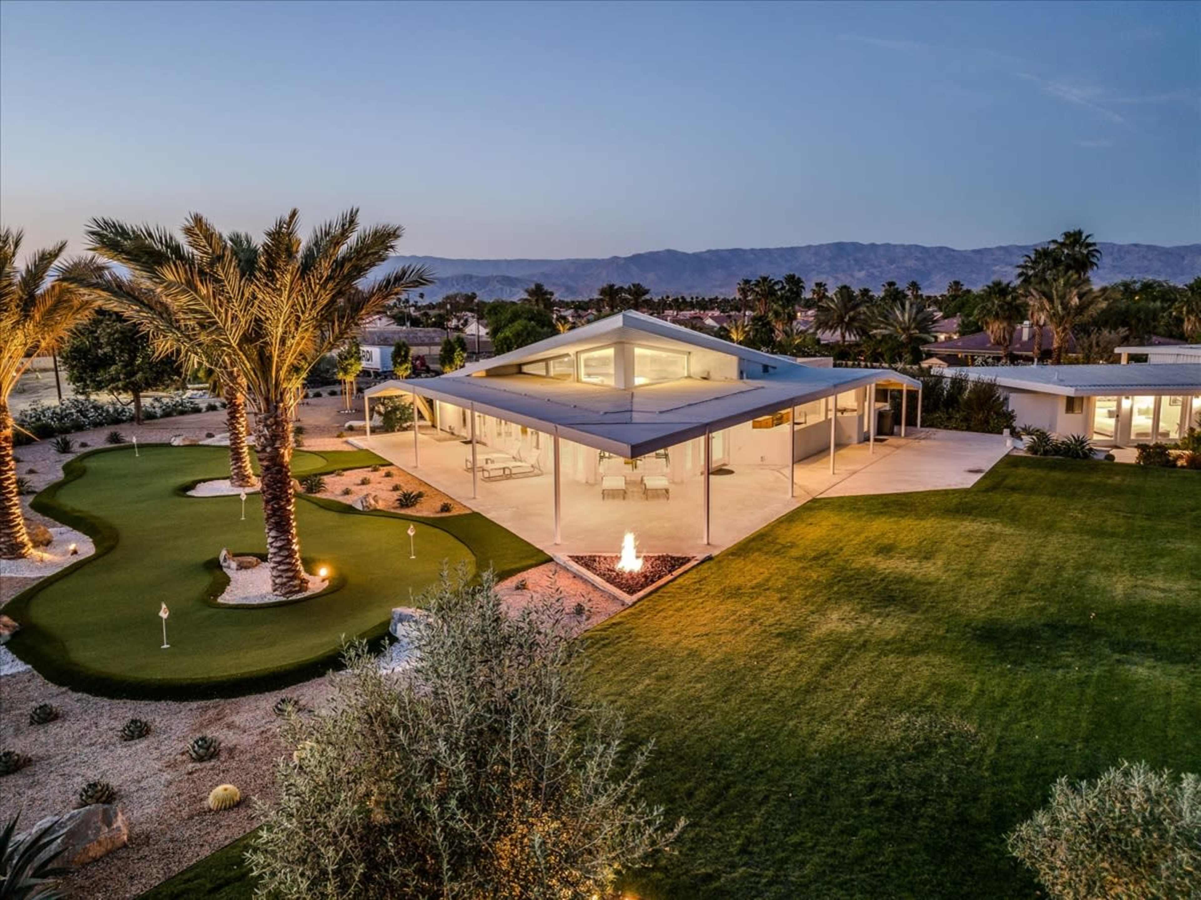 A modern house with a flat roof is situated amidst palm trees and landscaped green spaces, featuring an outdoor fire pit.