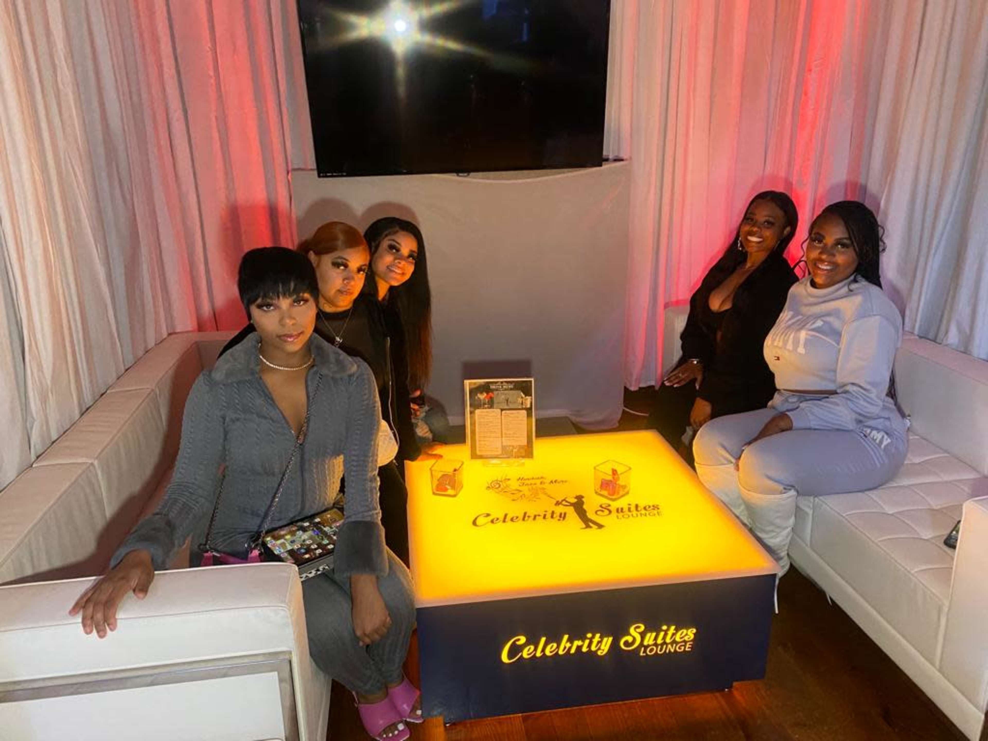 A group of five women poses together at a lounge, seated around a brightly lit table with the label "Celebrity Suites Lounge."