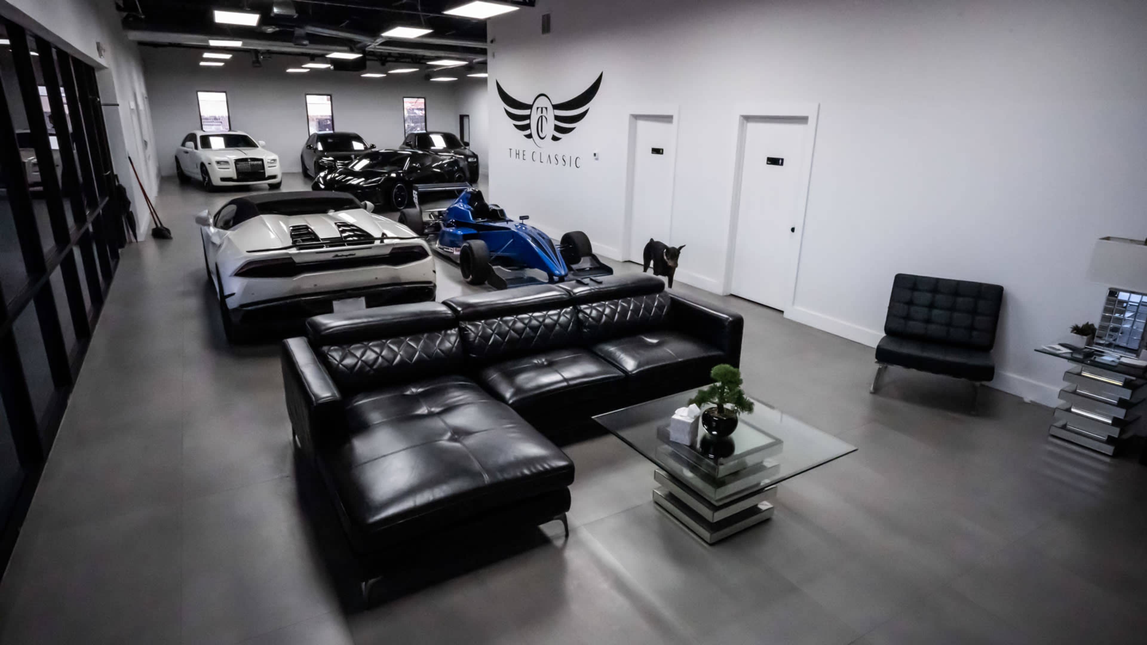 A modern showroom featuring several luxury cars, a black leather couch, a glass coffee table, and a sleek, minimalist design.