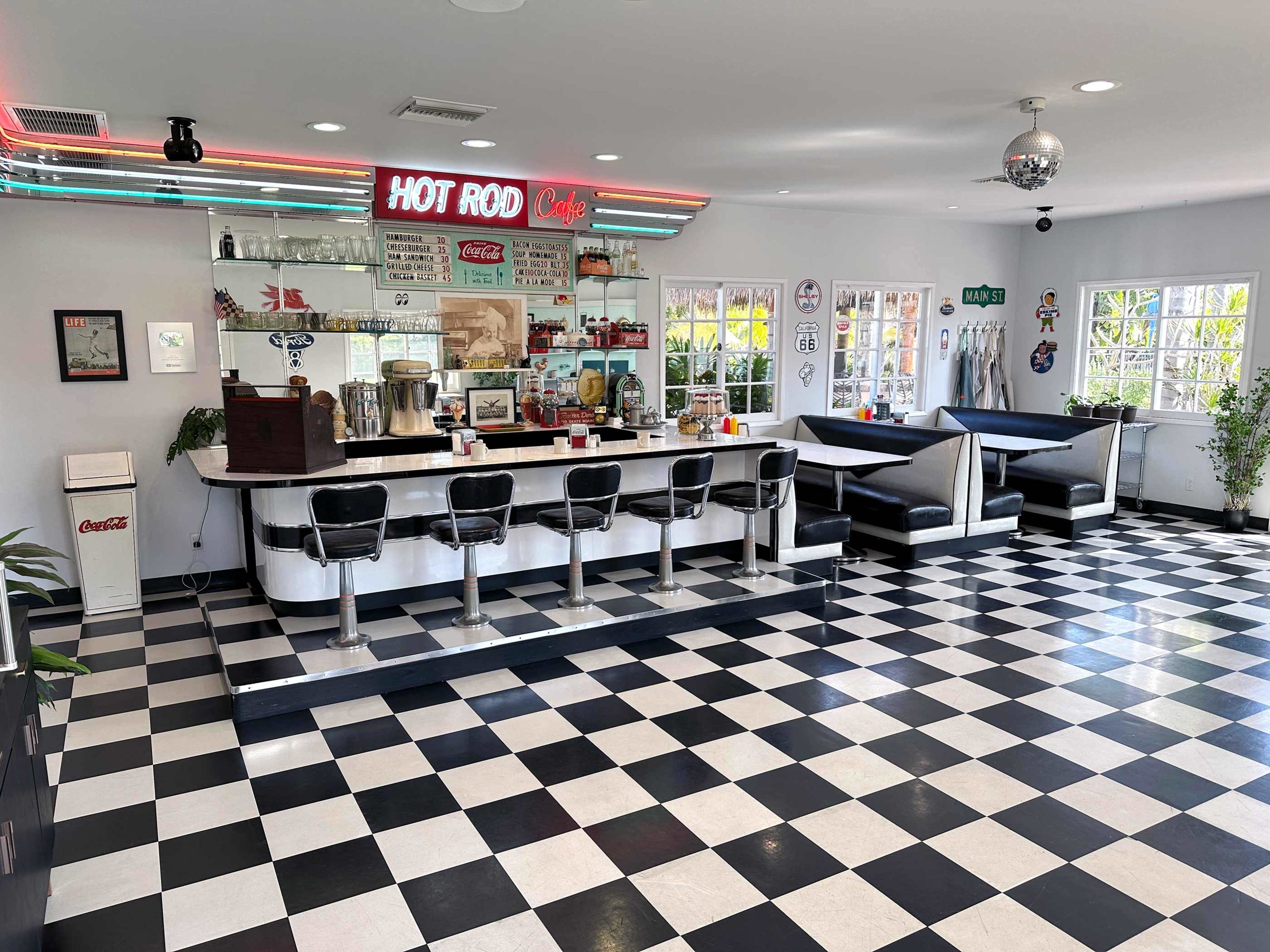 The image shows a vintage diner with a checkered black and white floor, a long counter with stools on one side, and booths on the other, featuring retro decor and a neon sign that reads "HOT ROD Cafe."