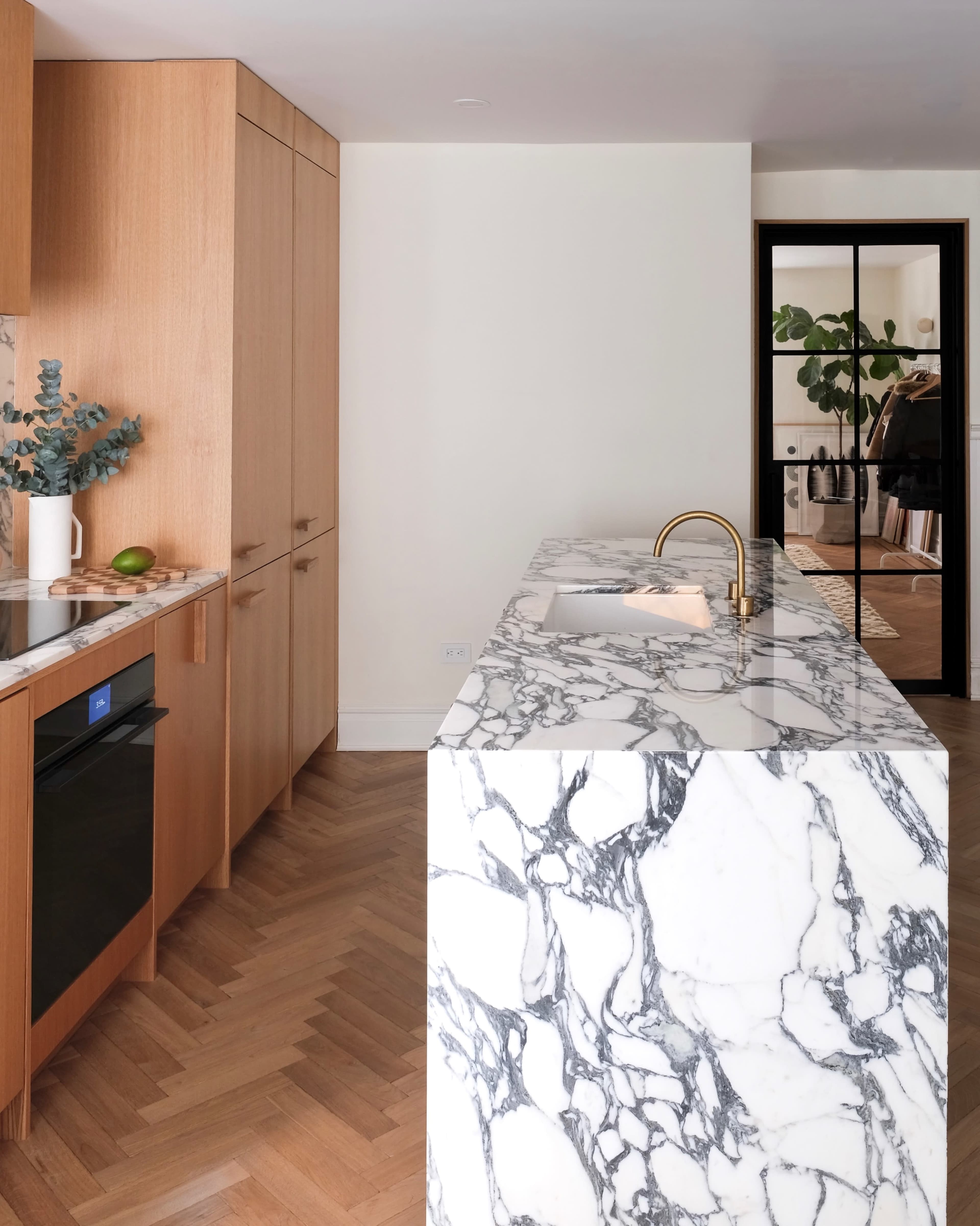 A modern kitchen features a marble island and wooden cabinetry against a backdrop of herringbone flooring.
