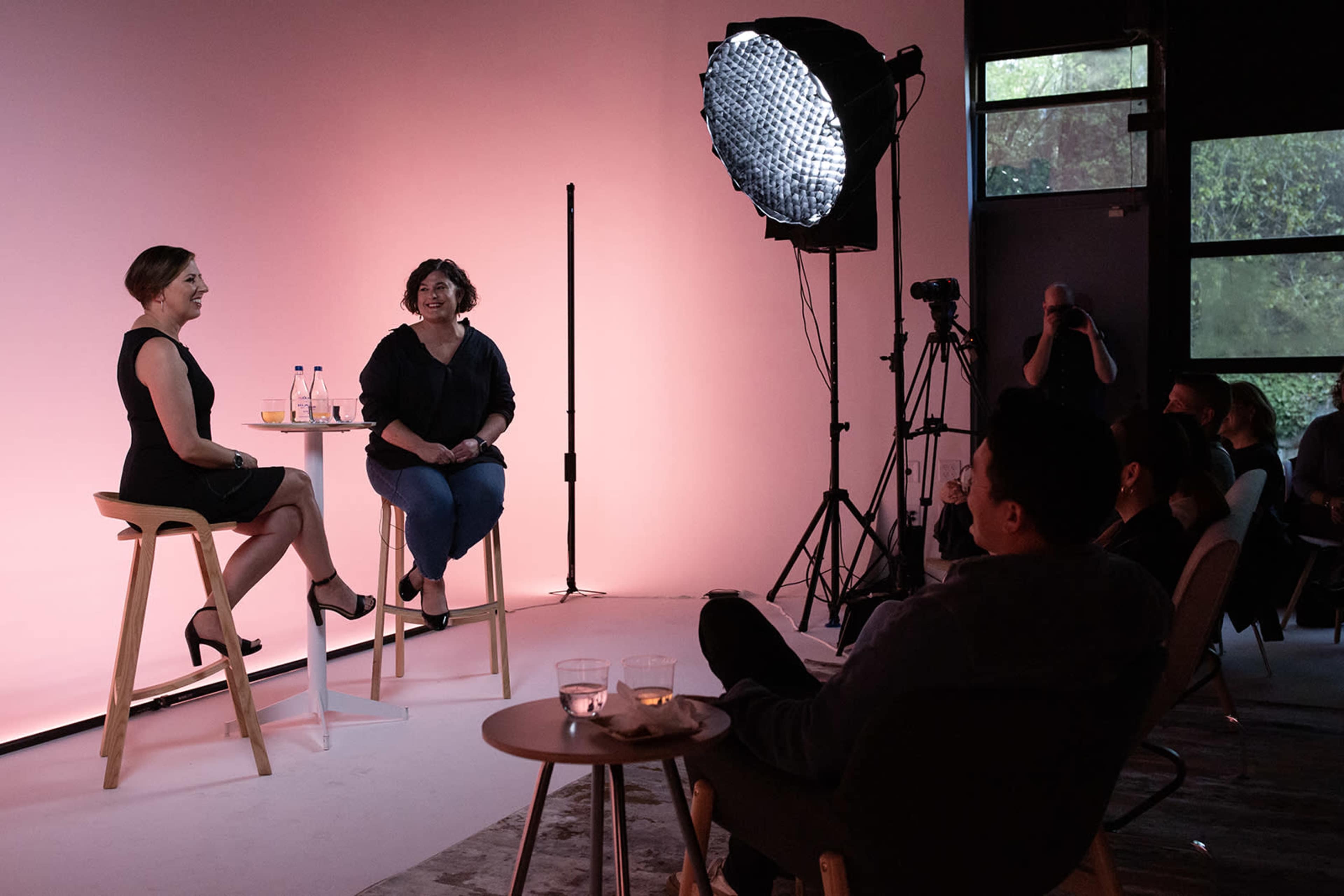 Two women sit on bar stools in front of a pink backdrop, engaging in conversation, while an audience observes in a studio setting.