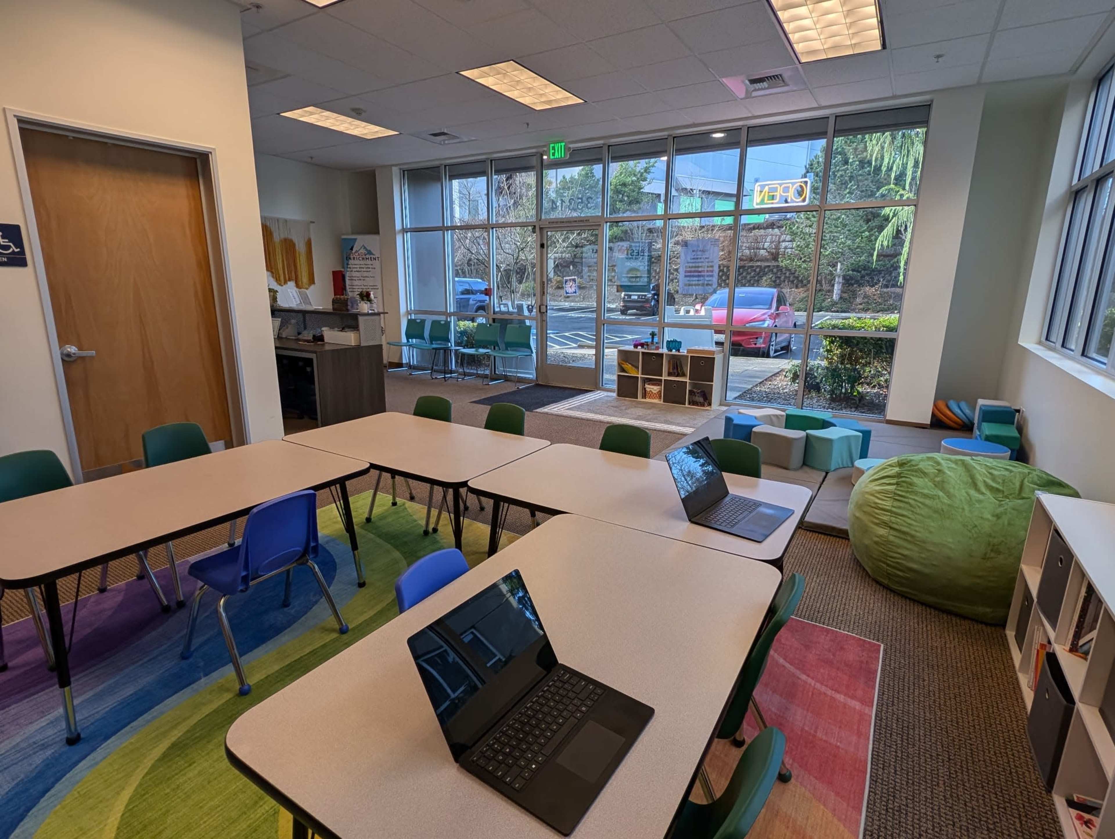 The image shows a classroom setting with multiple tables arranged, laptops open on the tables, and a seating area featuring a bean bag in a brightly lit environment with windows overlooking an outdoor space.