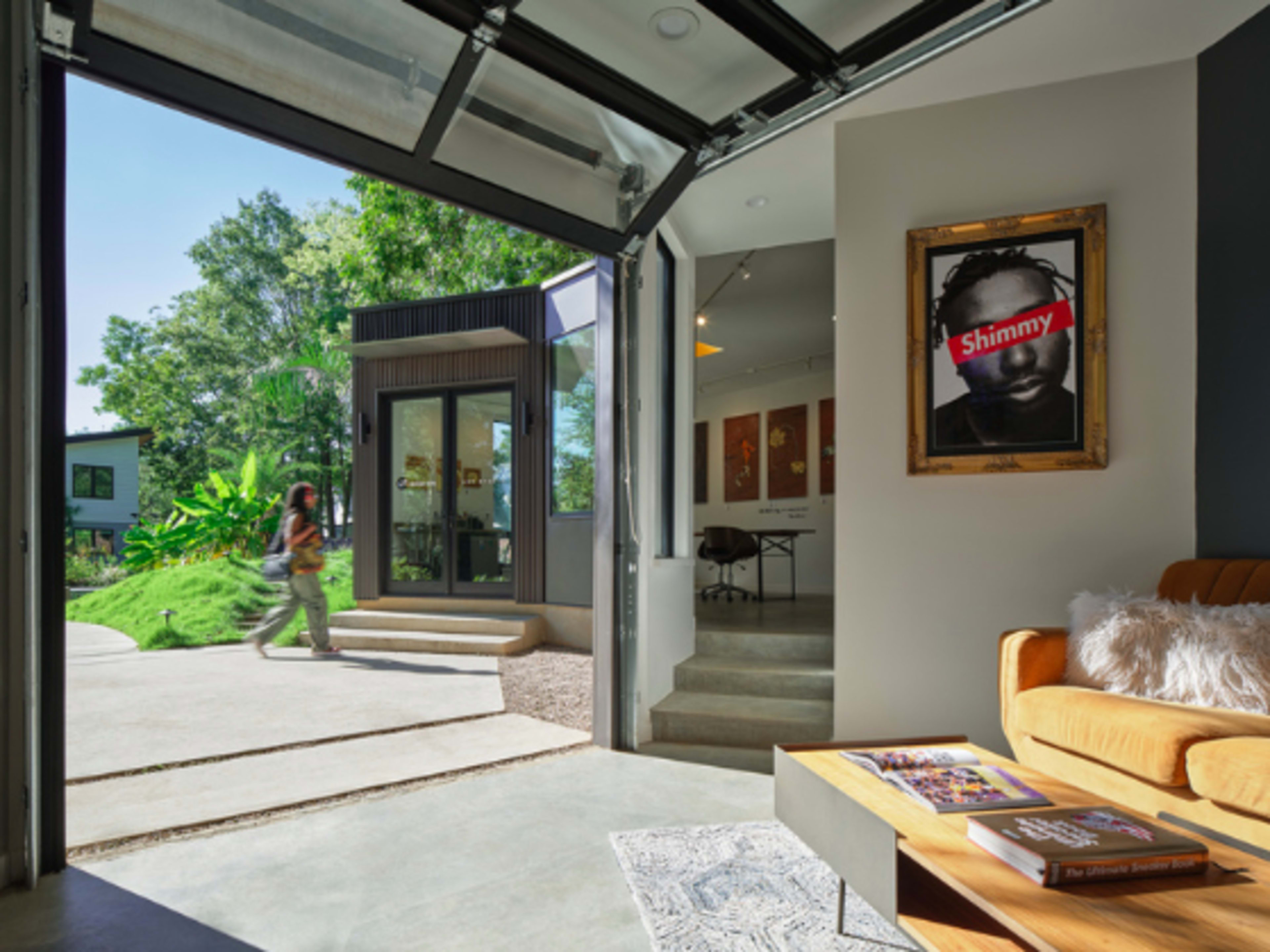 A person walks past an open garage door leading into a modern interior space featuring a framed artwork on the wall and a sofa.