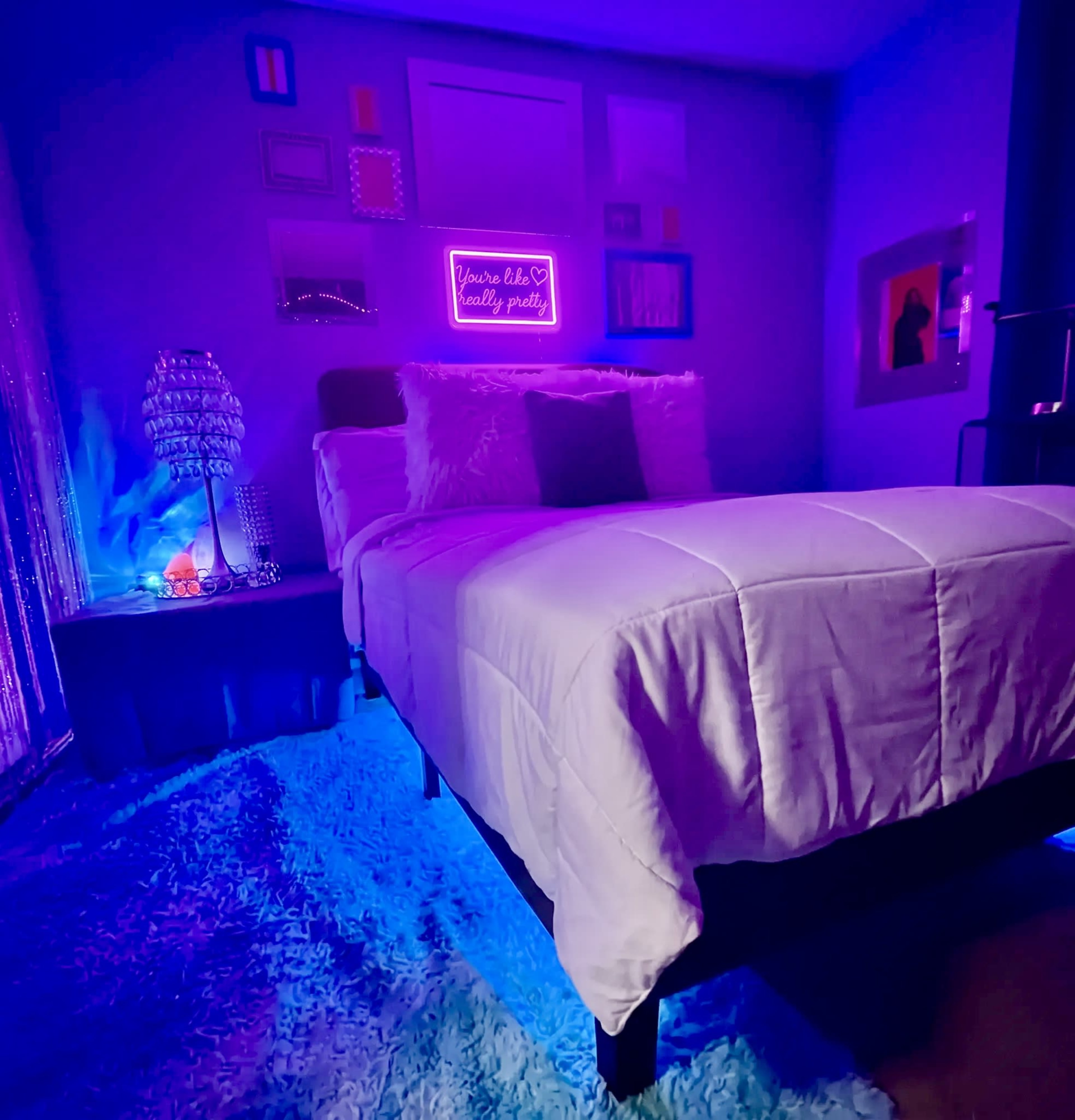 A neatly arranged bedroom features a bed with light bedding, a bedside table with a lamp, and colorful neon lighting illuminating the walls.