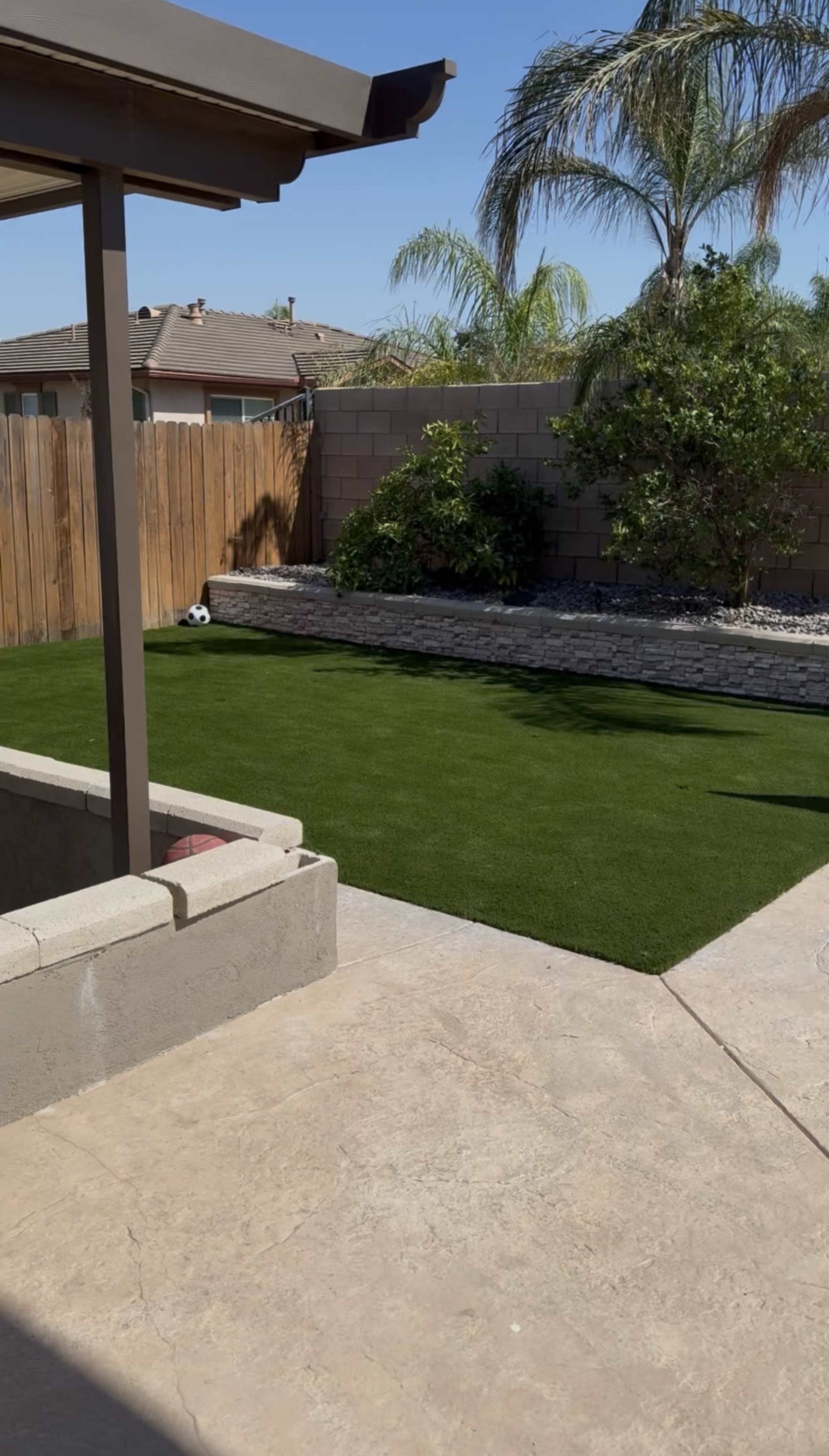 The image shows a backyard with a green artificial grass area bordered by a stone wall and surrounded by plants, with a concrete patio in the foreground.