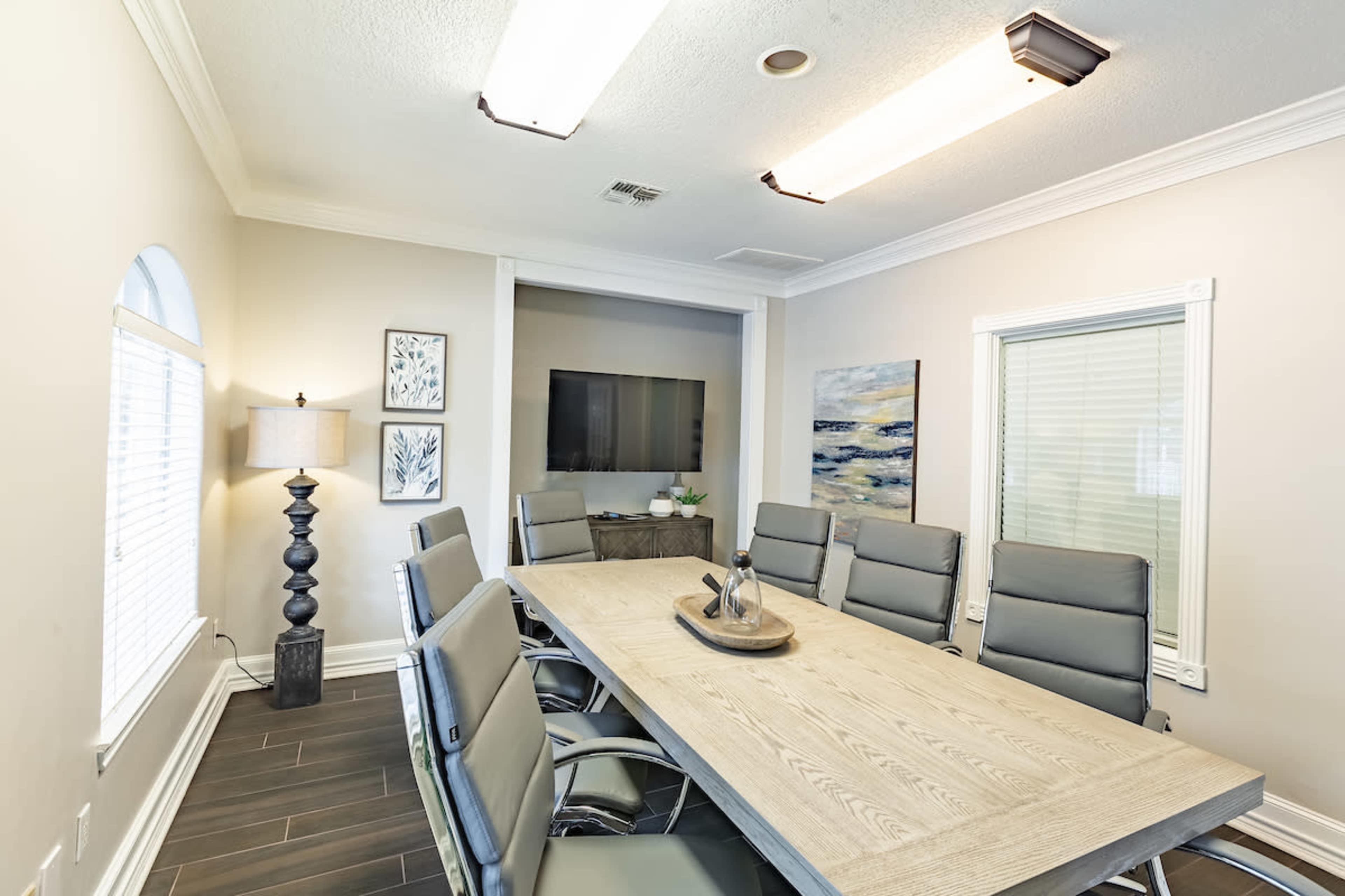 A modern conference room featuring a long wooden table surrounded by ergonomic chairs, with a television mounted on the wall and large windows allowing natural light.