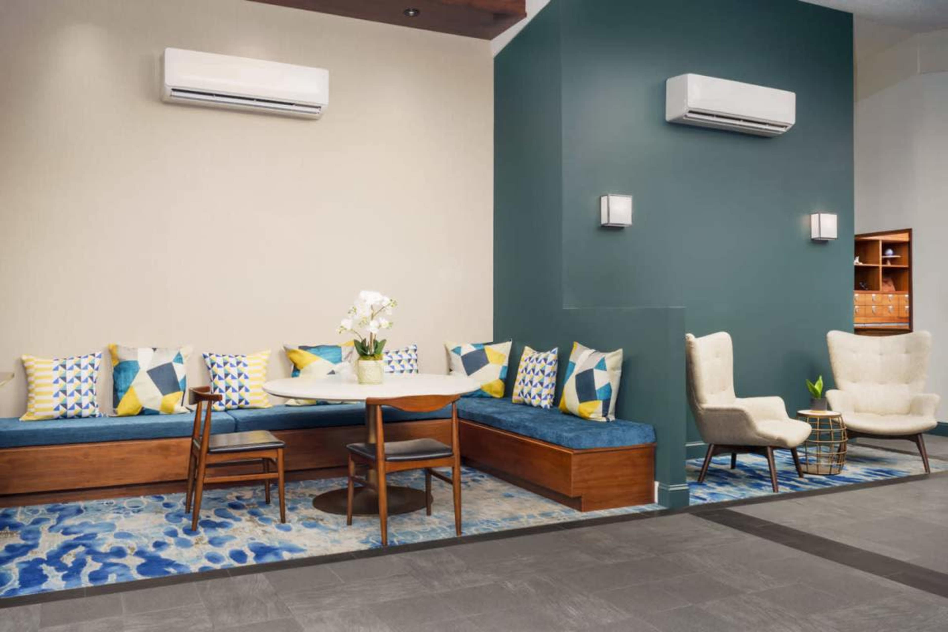 The image shows a modern lounge area featuring a blue and white patterned rug, a wooden seating space with colorful pillows, and two upholstered chairs near a wall.