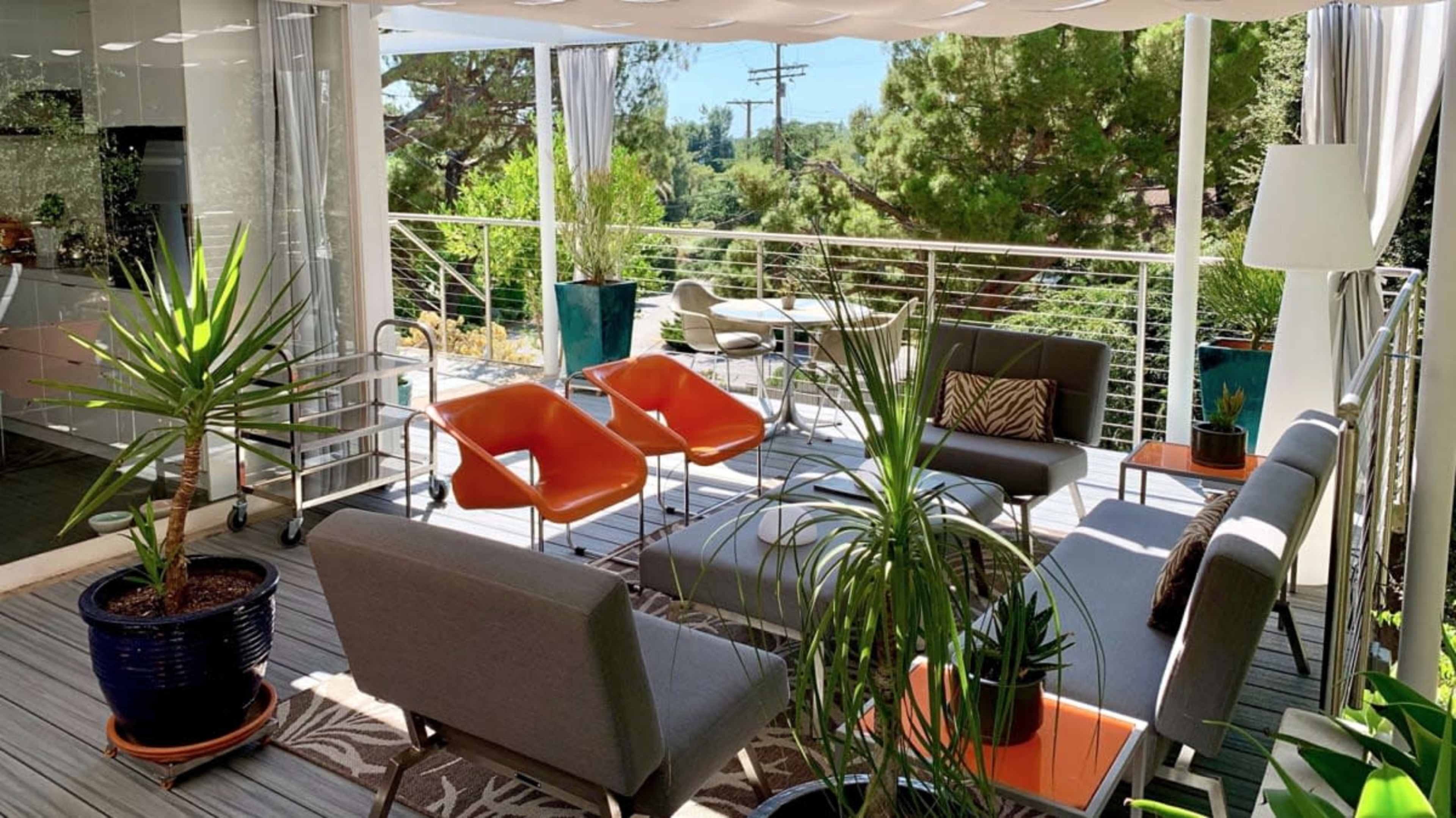 The image shows a modern outdoor patio with sleek furniture, potted plants, and a view of greenery in the background.