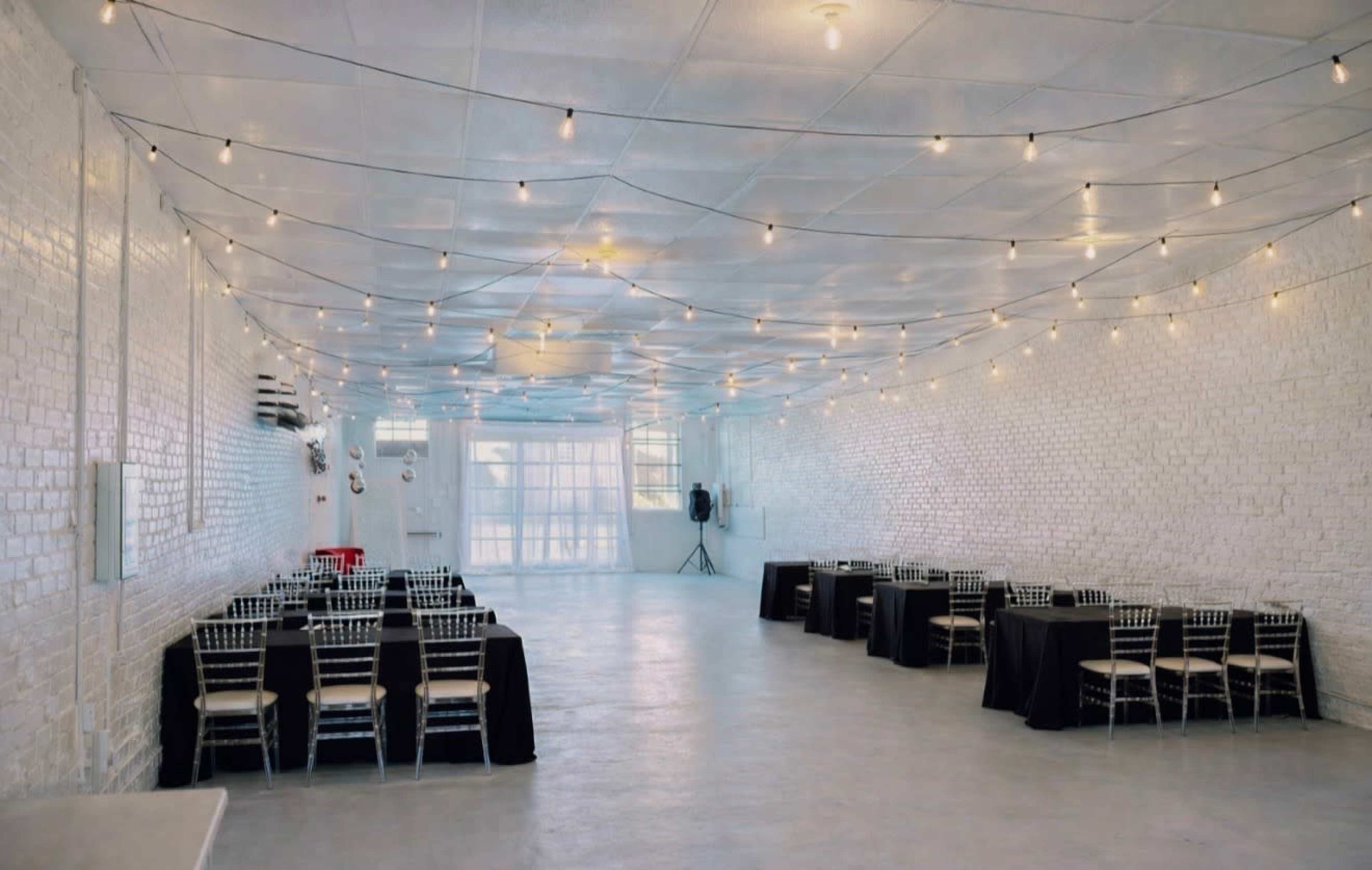 The image shows a spacious, empty event hall with white brick walls, string lights overhead, and rows of black tables set with chairs.