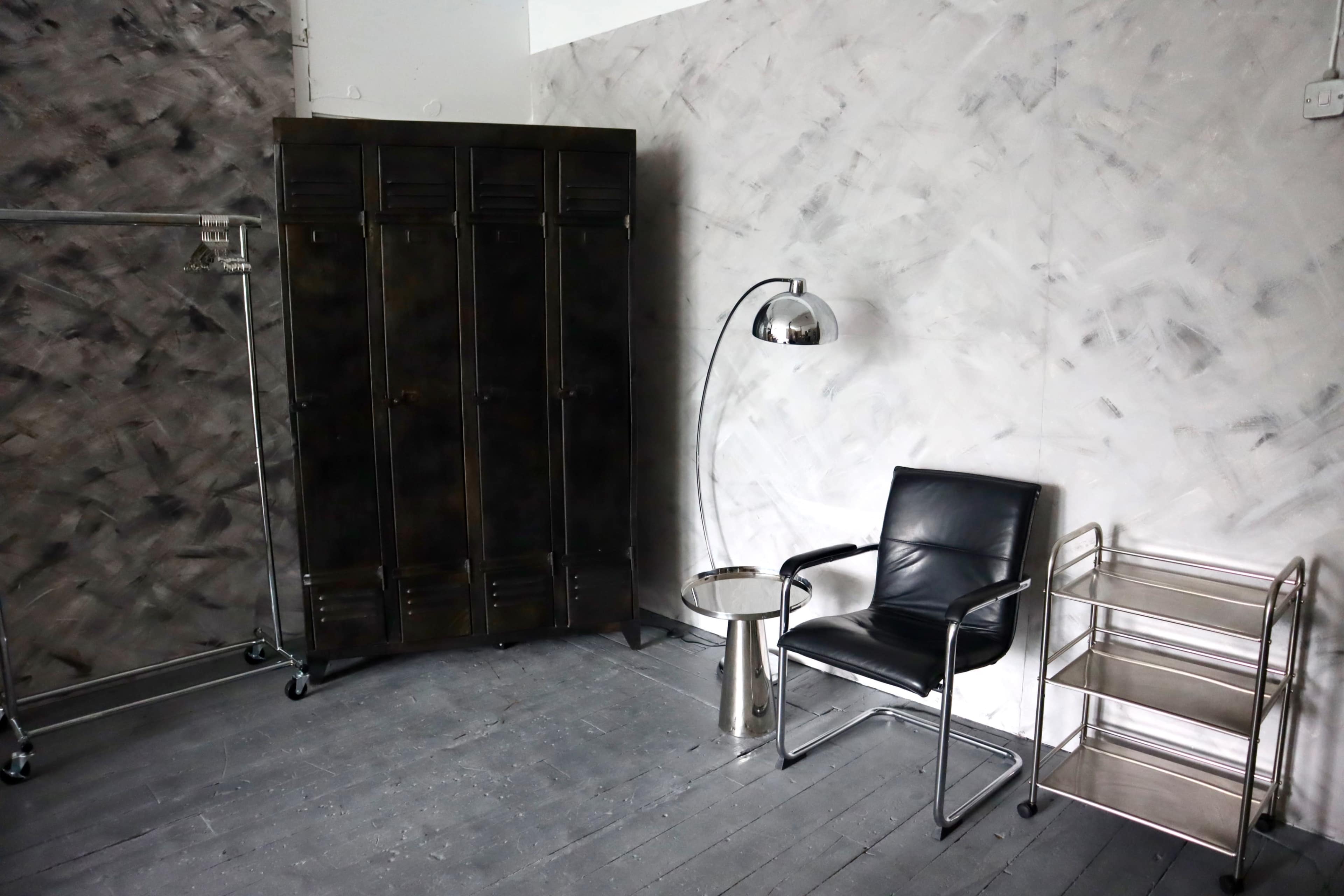 The image shows a minimalist interior space featuring a black chair, a metallic lamp, a storage locker, and a small side table on a wooden floor.