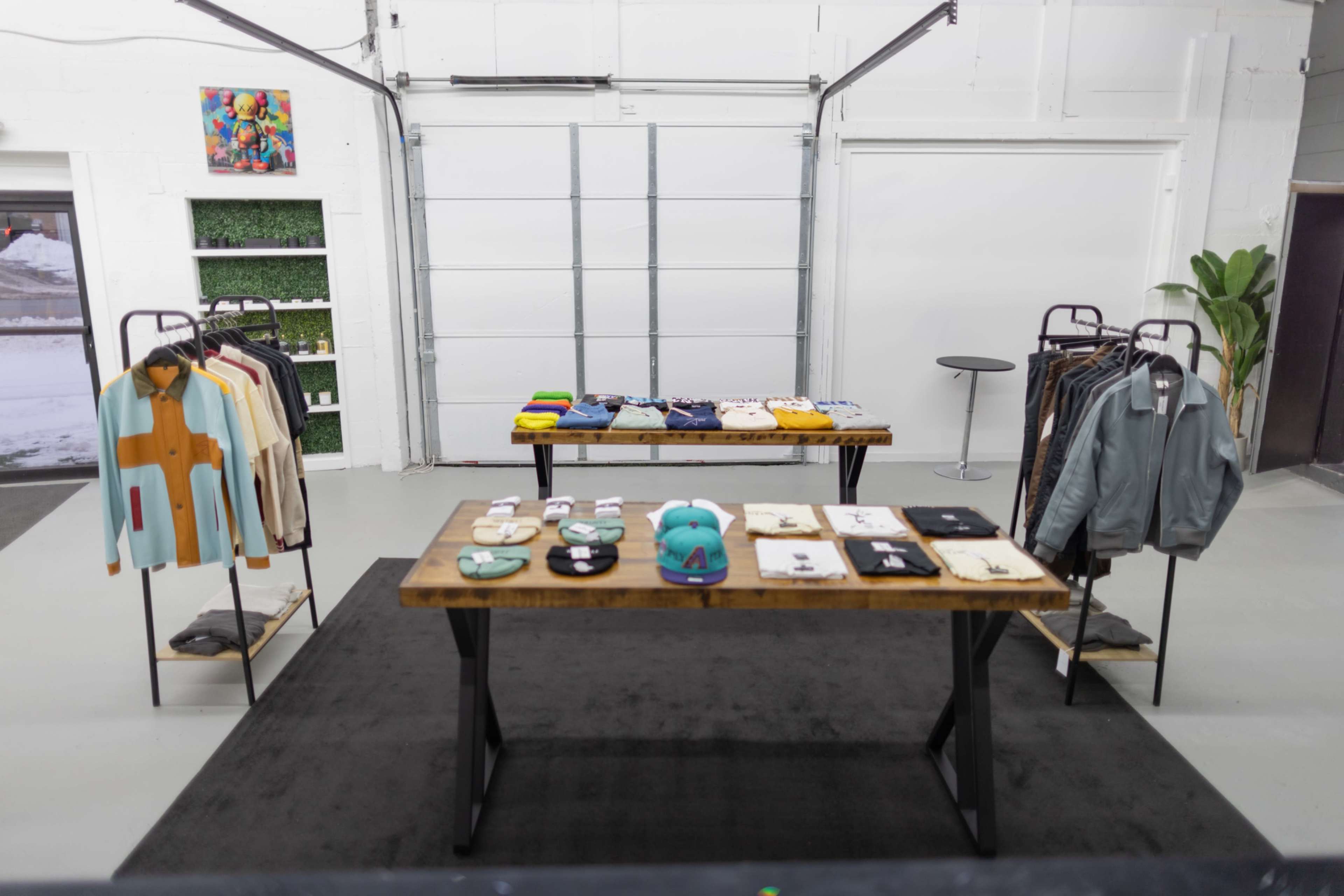 The image shows a modern clothing retail space with two wooden display tables featuring various apparel and accessories, set against a bright, minimally decorated backdrop.