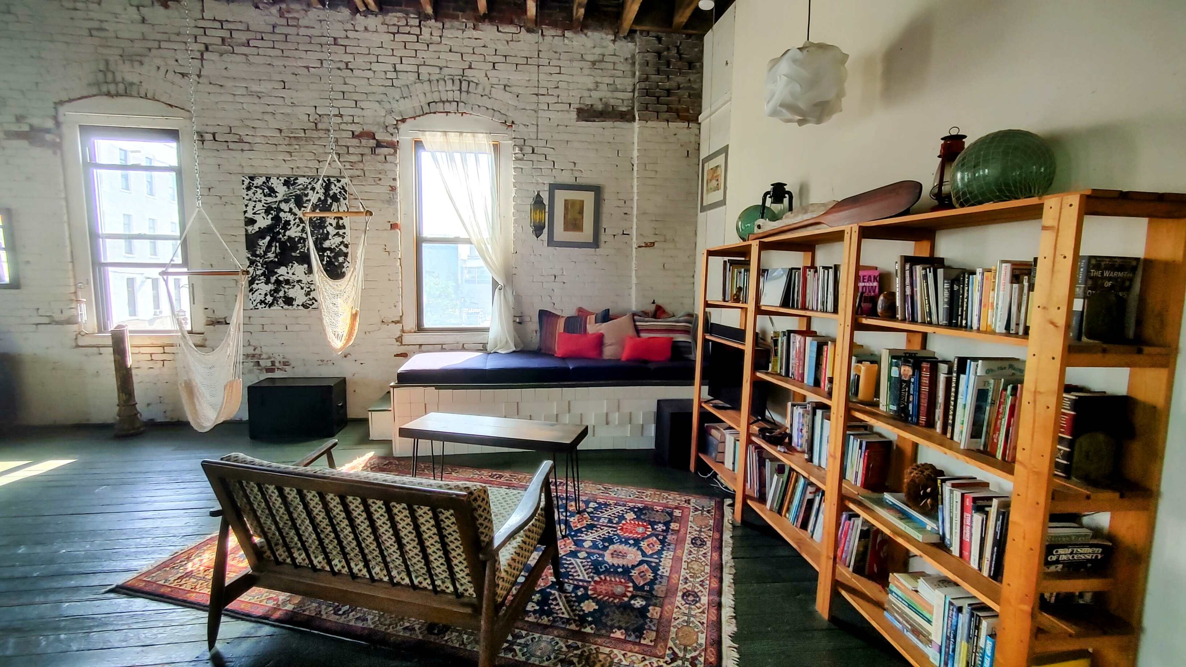 The image depicts a spacious room with exposed brick walls, a large bookshelf filled with books, a seating area with a patterned rug, and large windows allowing natural light to enter.