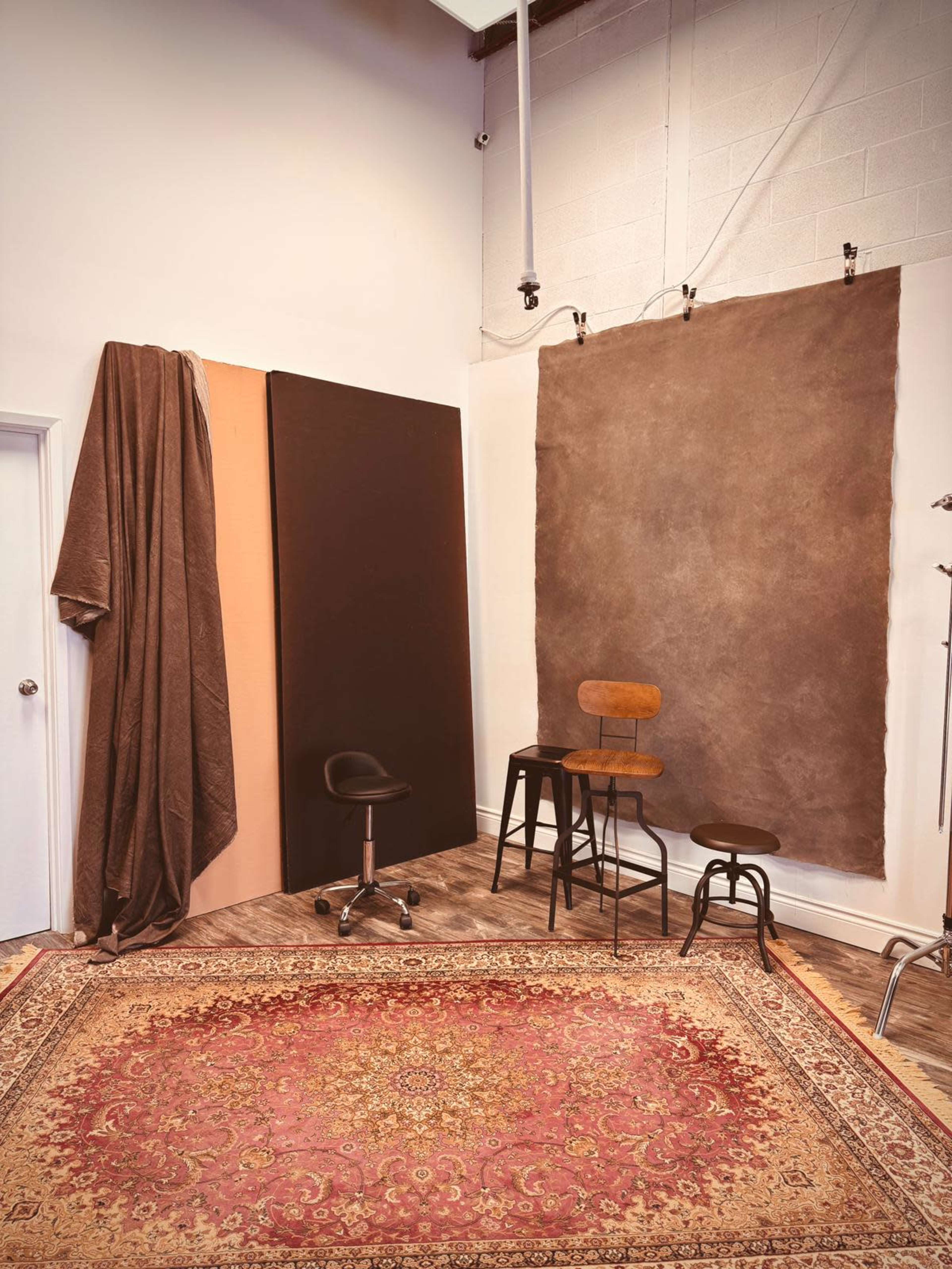 The image depicts a studio space with a patterned rug, two wooden stools, a rolling chair, and three backdrop panels in neutral colors, along with a draped fabric hanging on one side.