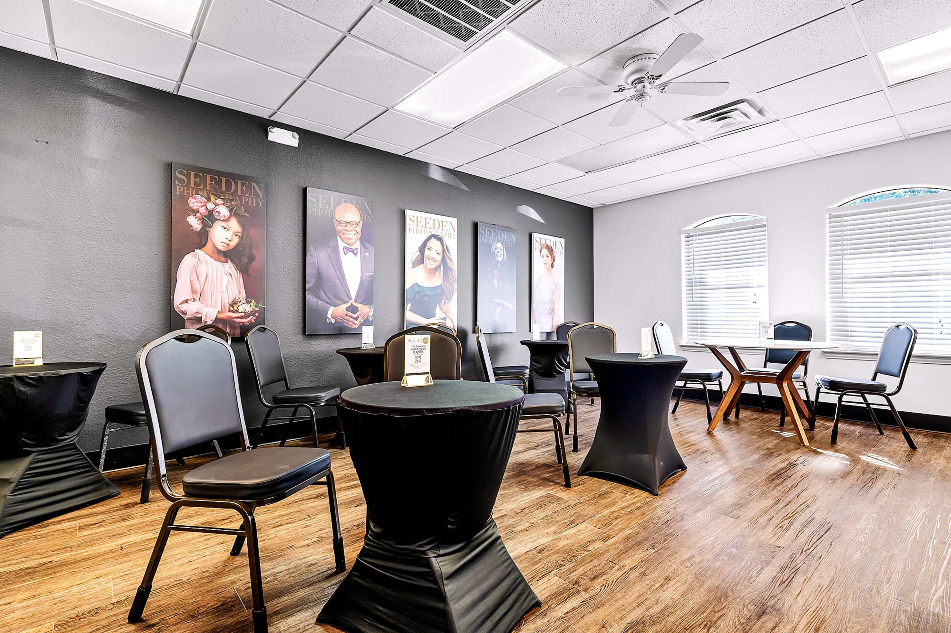 A room with black chairs and tables draped in black cloth, featuring large framed portraits on the walls and large windows allowing natural light.