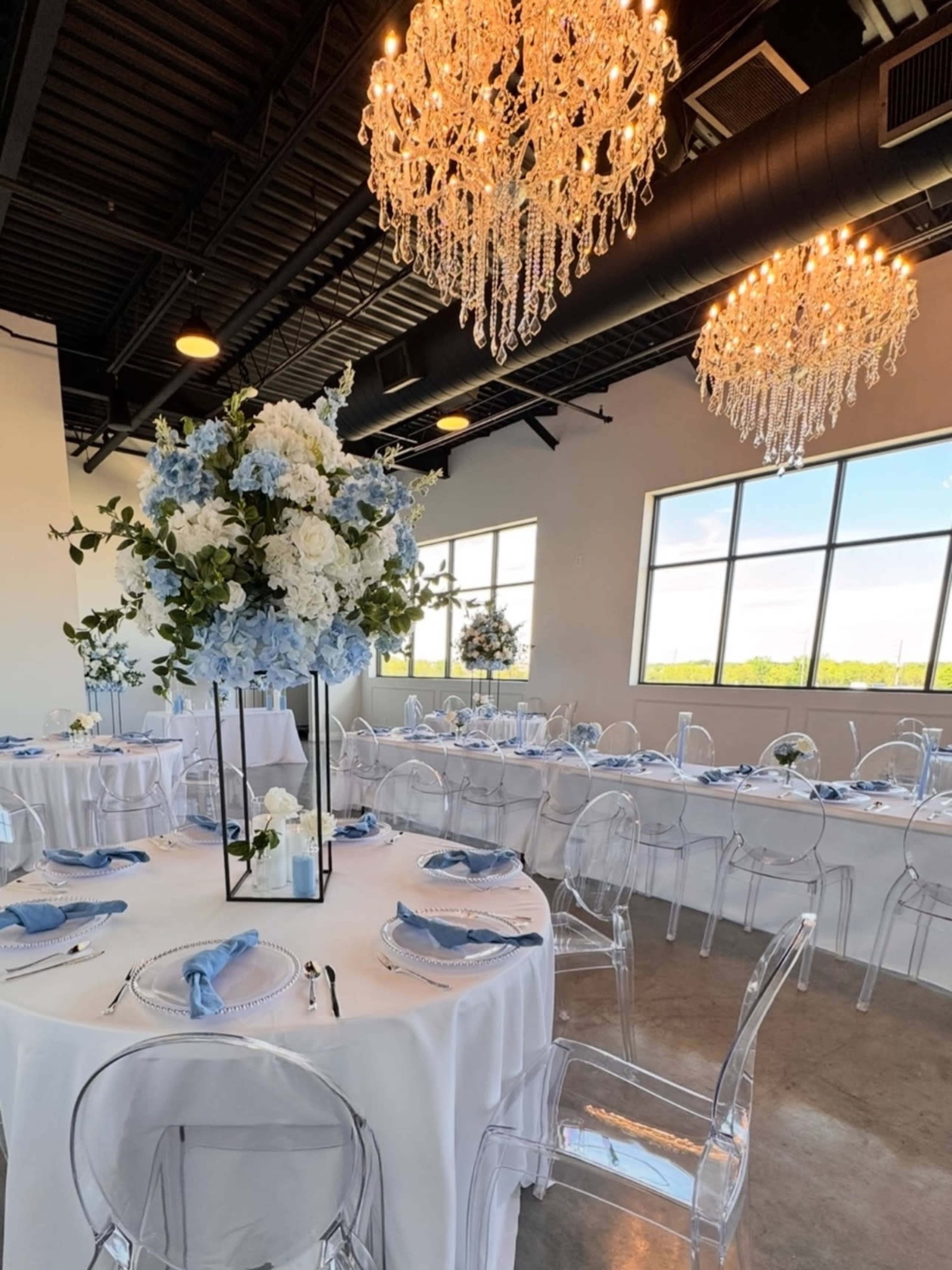The image shows a modern dining area with round tables dressed in white tablecloths, each featuring tall floral centerpieces and crystal chandeliers hanging from the ceiling.
