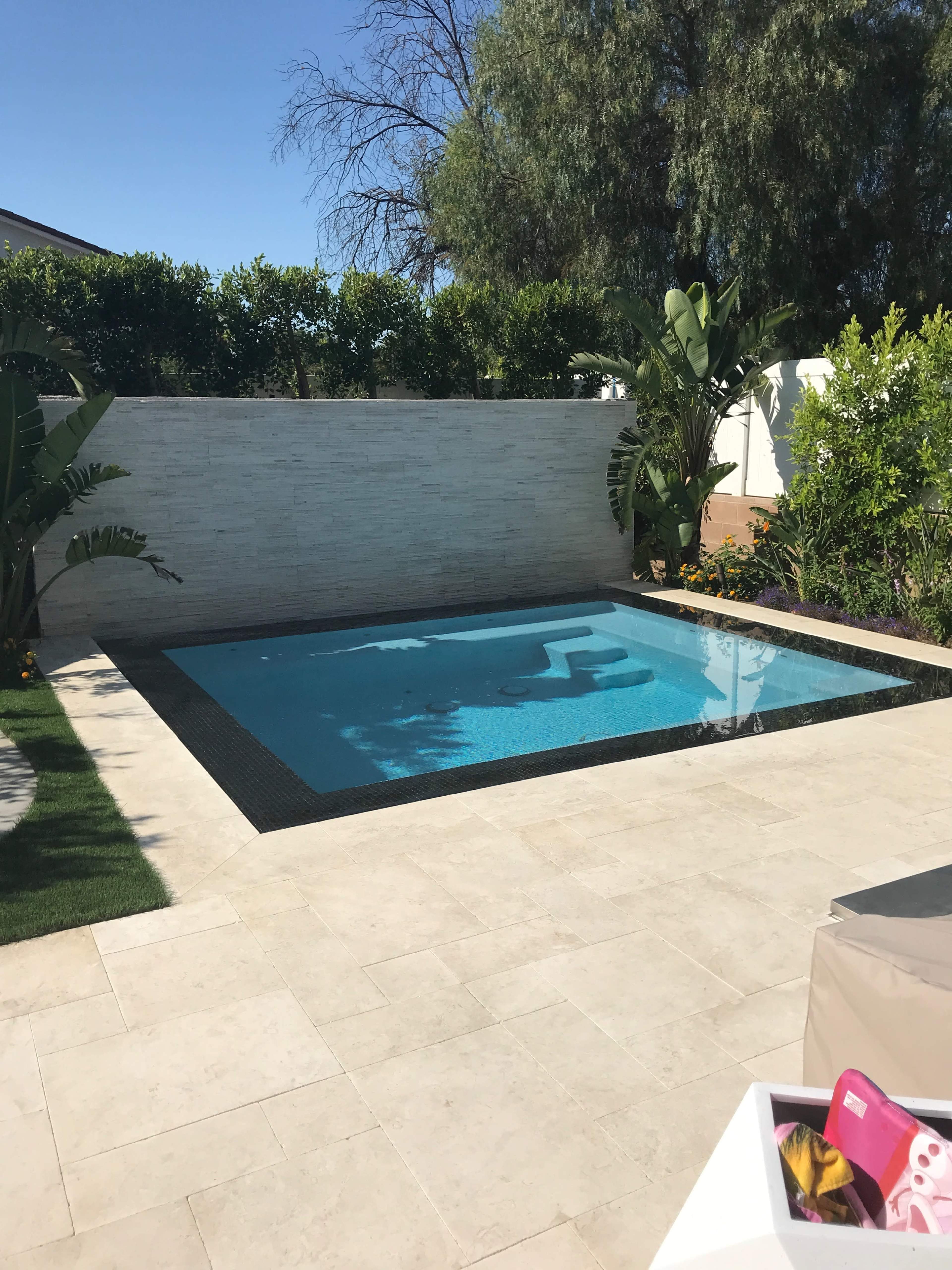 A rectangular swimming pool is surrounded by tiled flooring and greenery, with a white wall in the background.