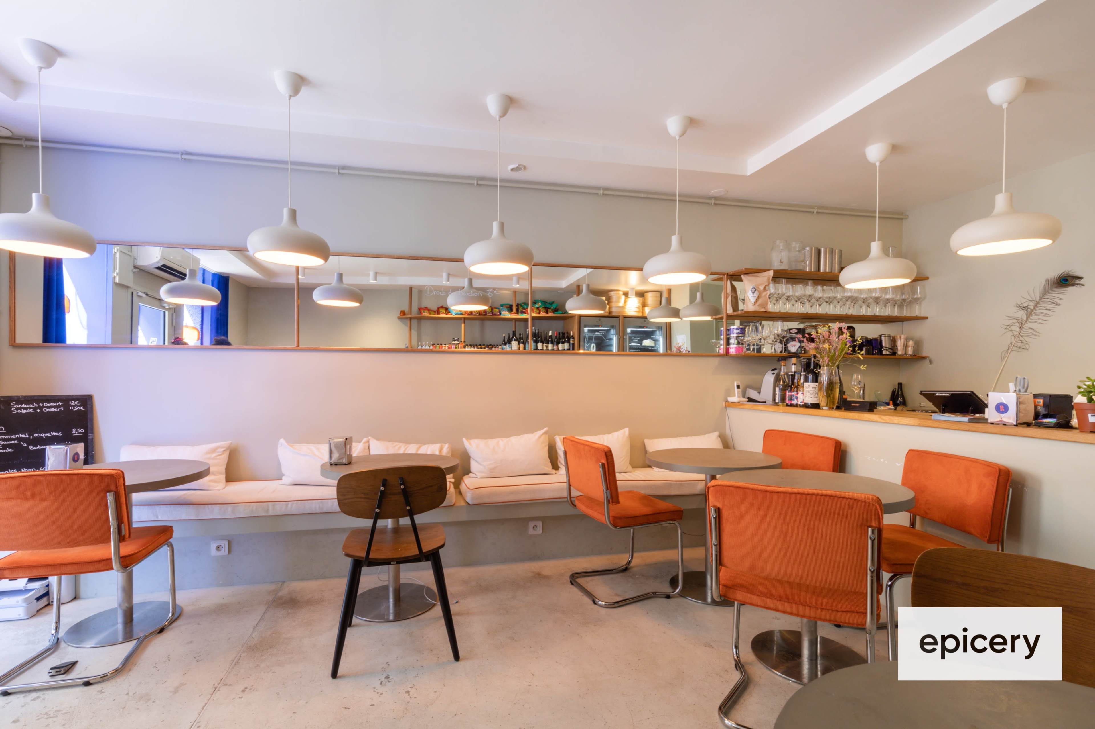 The image shows a modern café with round tables, orange cushioned chairs, and pendant lights hanging from the ceiling.