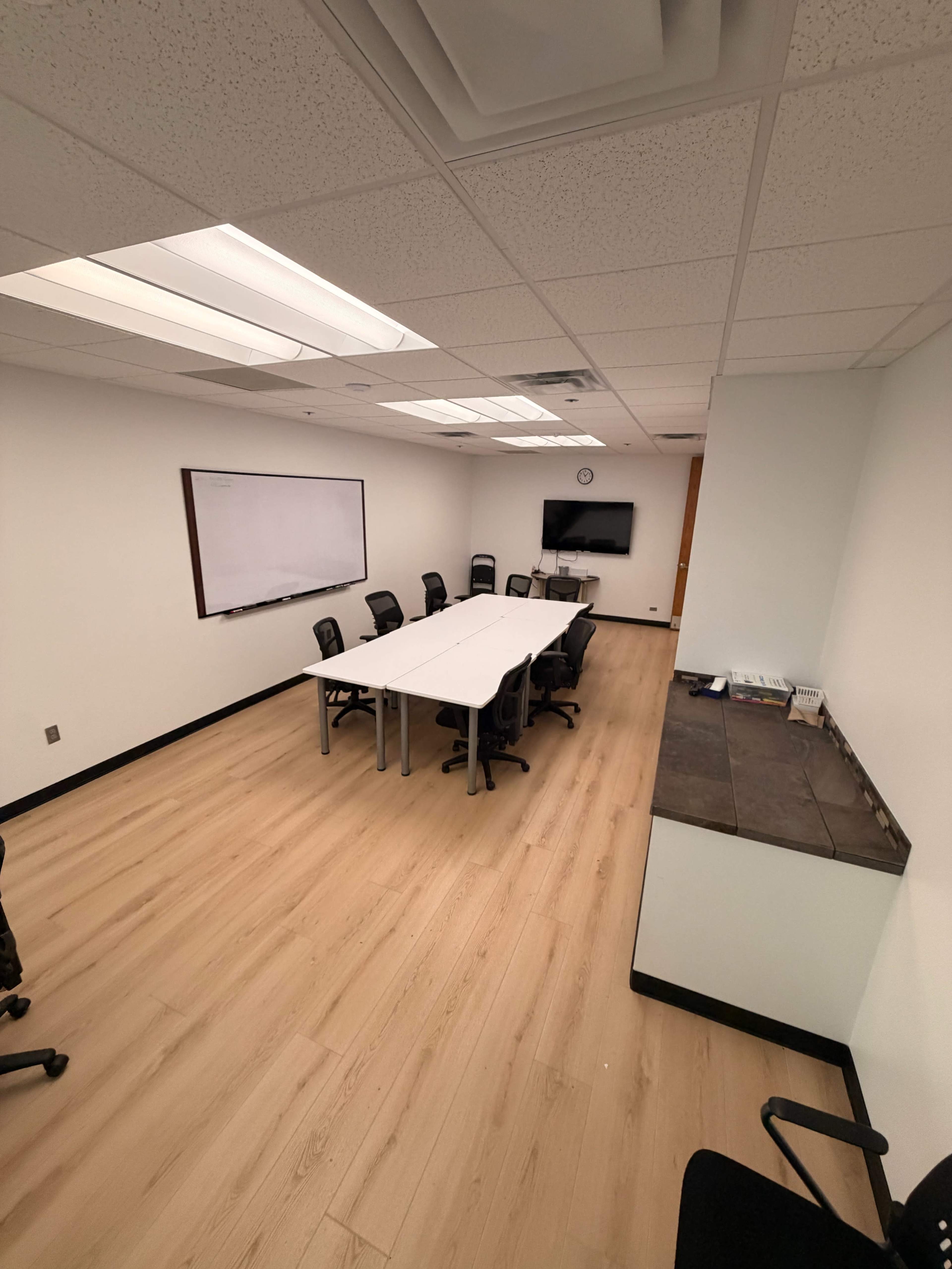The image shows a conference room with a long table surrounded by black chairs, a whiteboard on one wall, and a TV mounted on another wall.