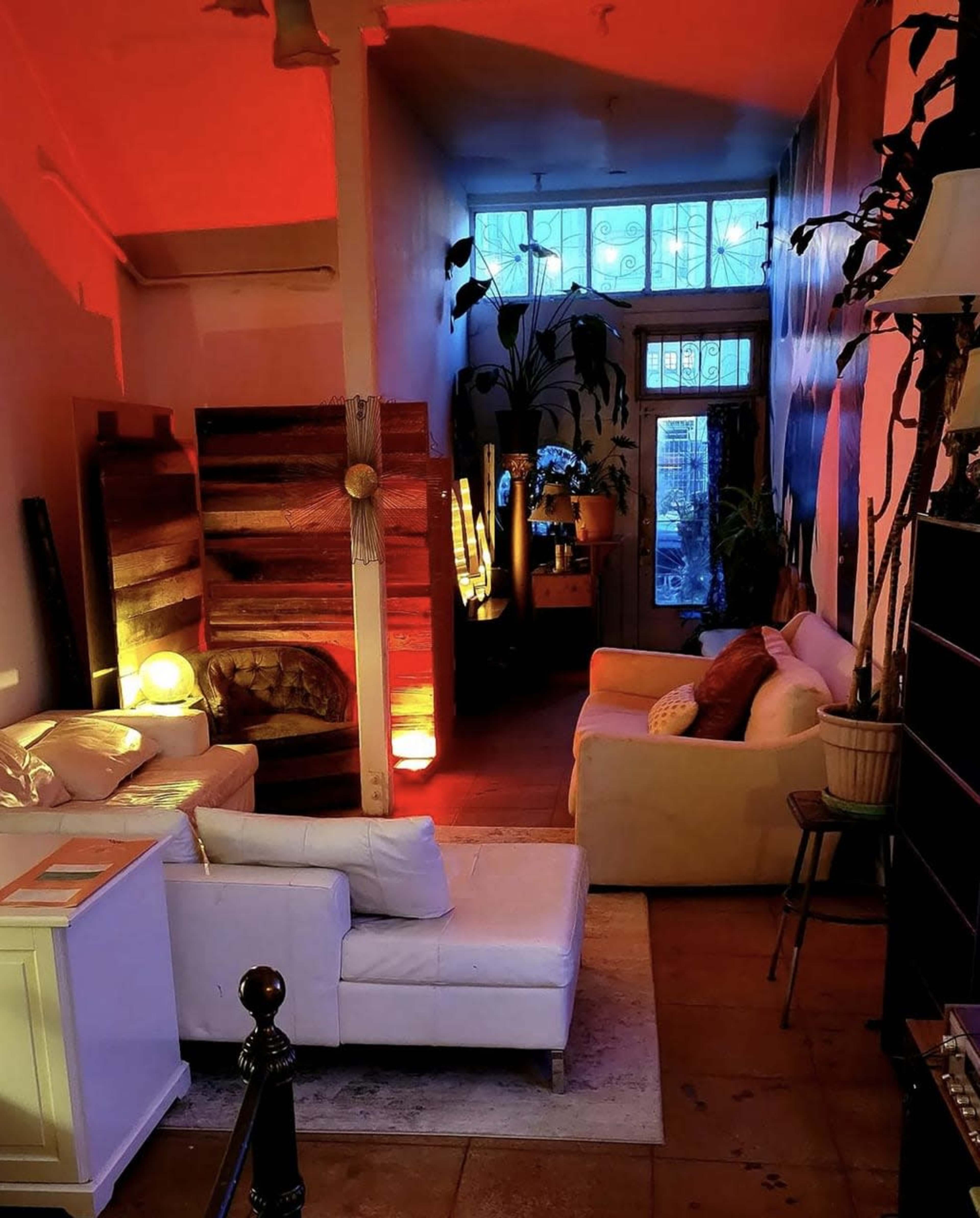 The image shows a cozy interior space with contrasting lighting, featuring a seating area with couches and plants, leading towards a brightly lit room at the back.
