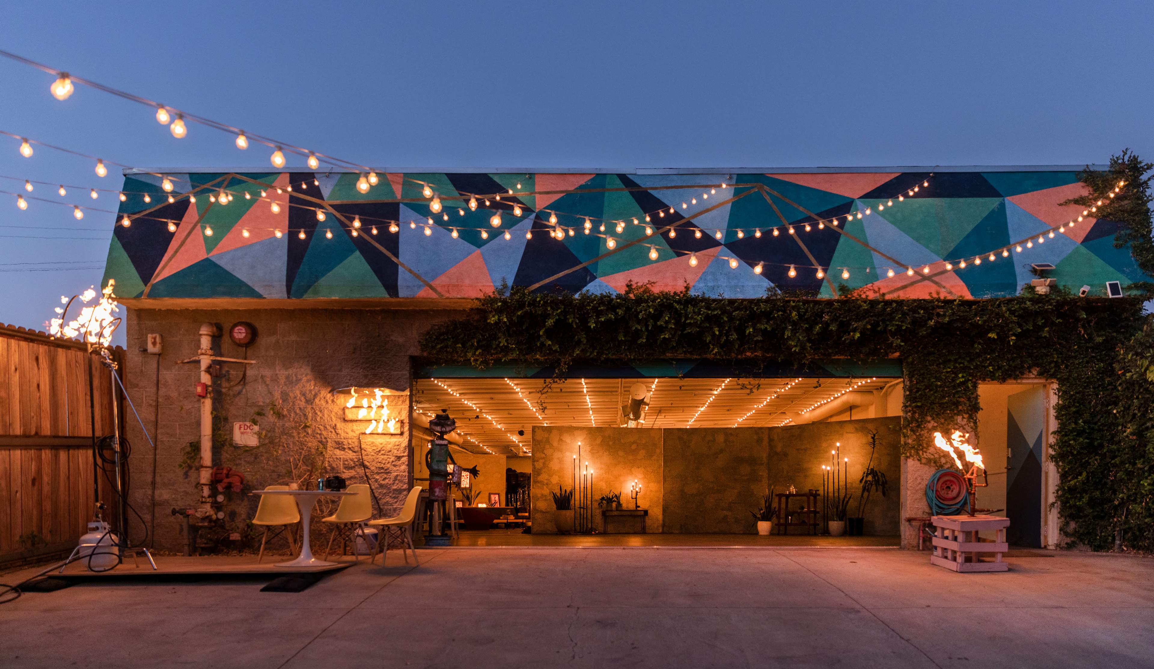 The image shows a modern building with a colorful geometric mural and string lights, featuring an open garage space with shelves and decor inside.