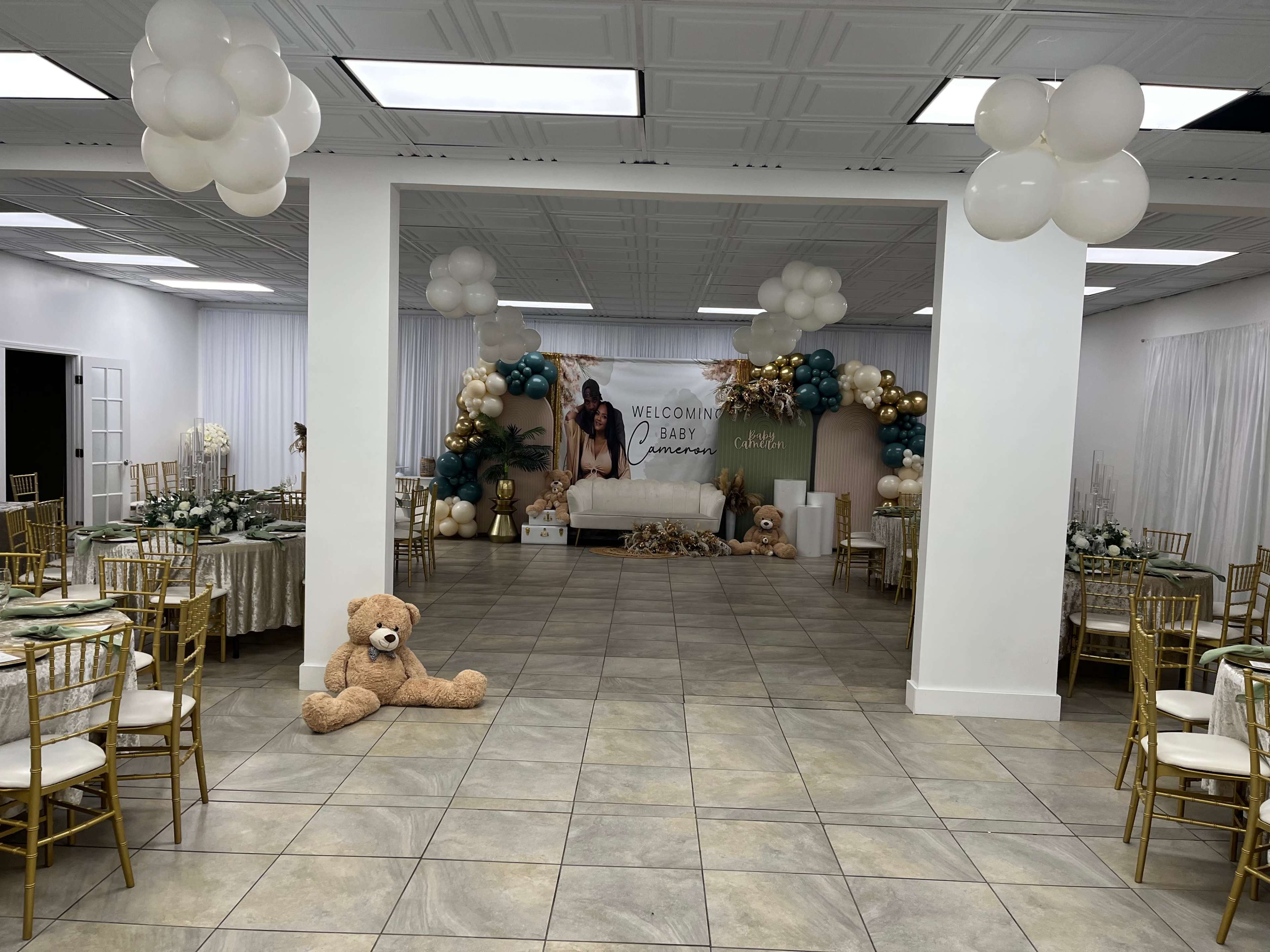 A decorated event space set up for a baby shower, featuring tables with gold chairs, a sofa adorned with balloons, and a large banner welcoming the baby.