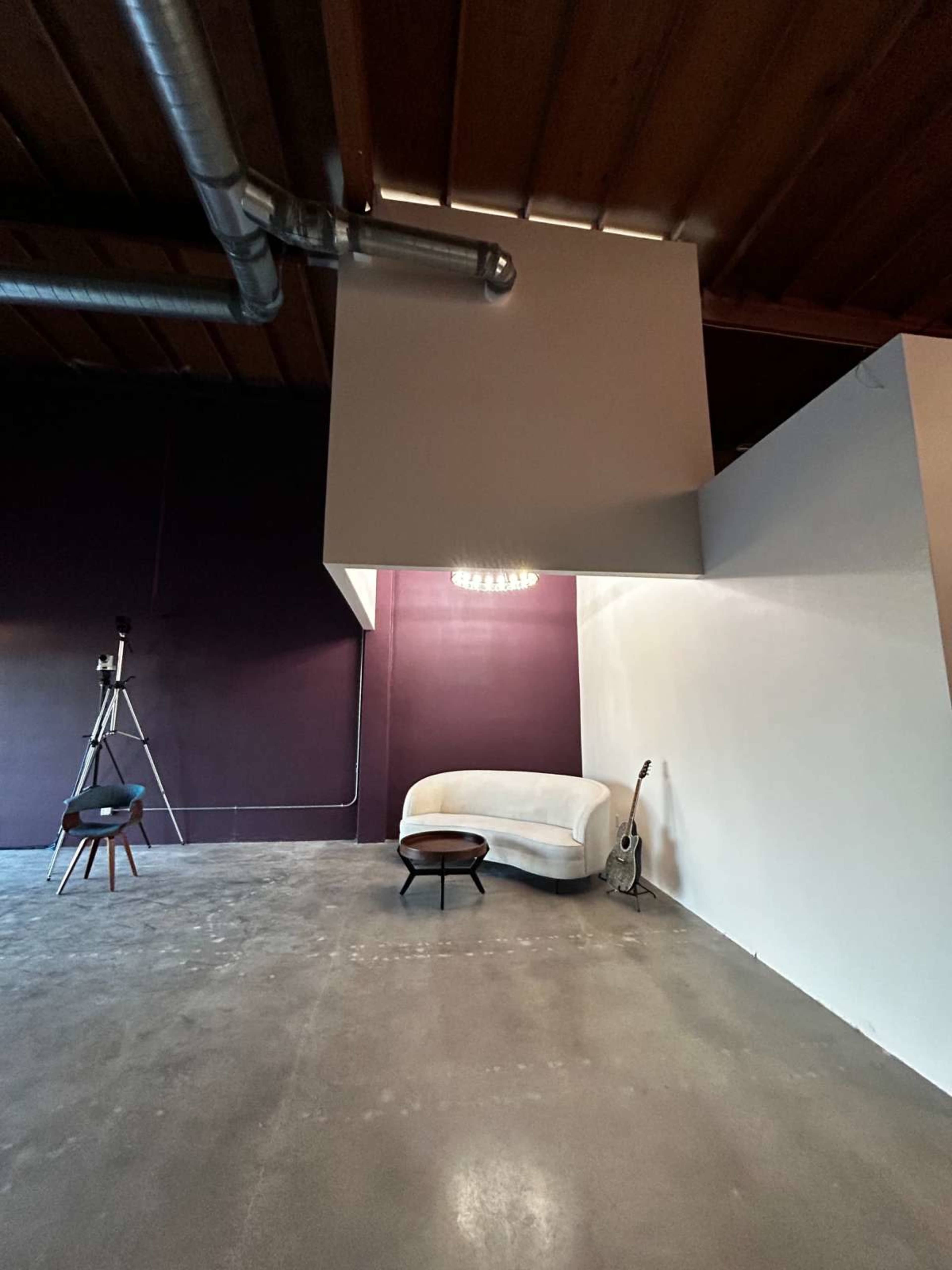 The image shows a spacious room with a light-colored couch, a guitar leaning against a wall, a small round table, and a tripod with a camera in one corner.