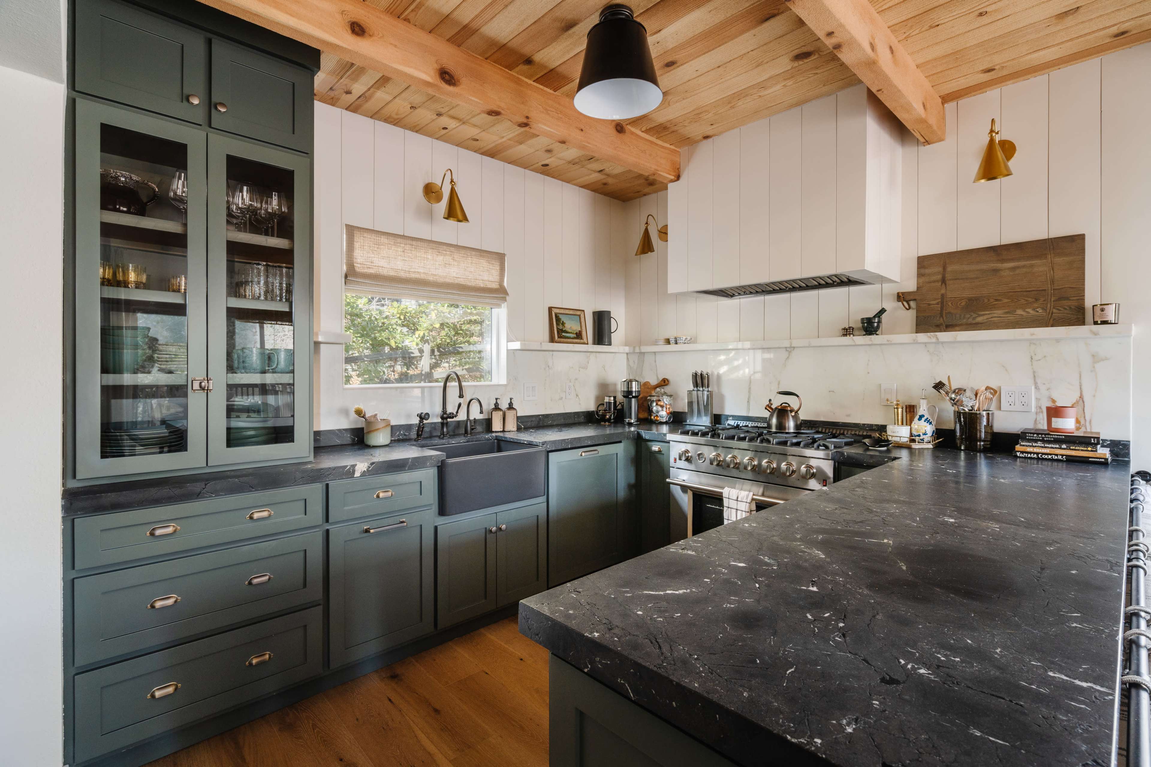 A modern kitchen with green cabinetry, a large black marble countertop, and wooden beams on the ceiling.