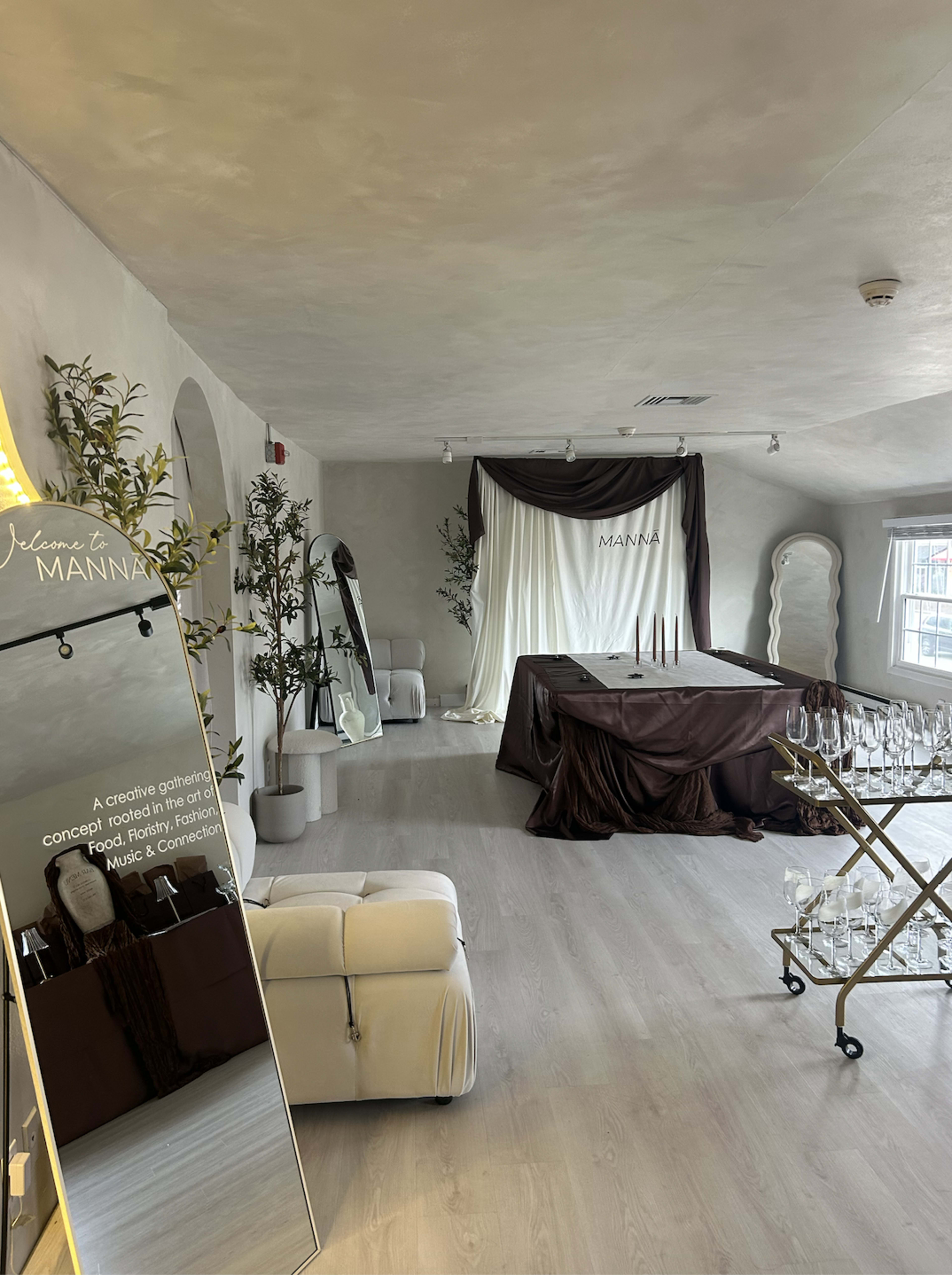 The image shows a spacious, elegantly decorated room with light walls, a seating area, a large table covered in rich brown fabric, and a mirrored cart with glasses.