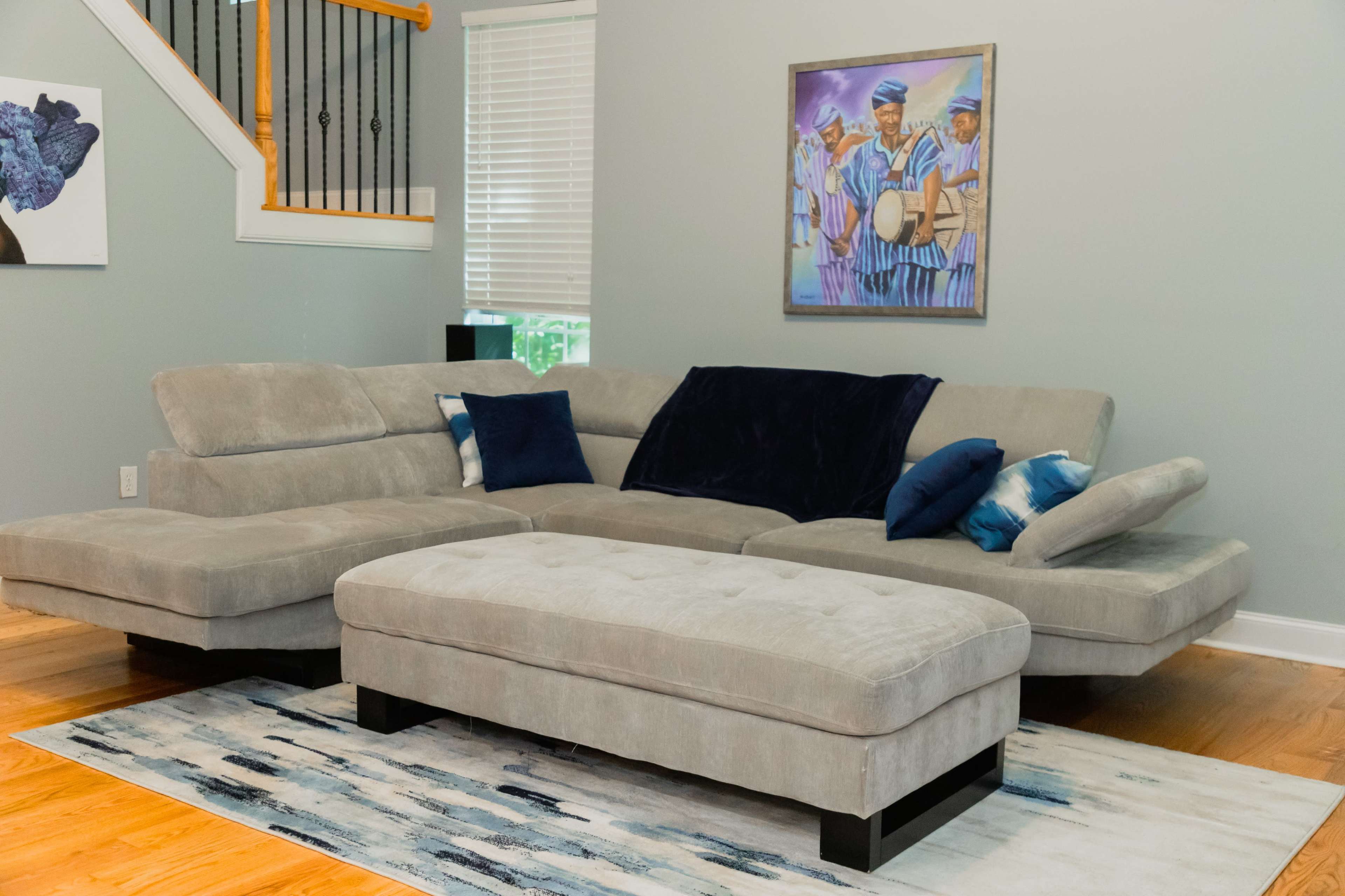 A gray sectional sofa with navy blue and patterned pillows sits on a blue and white area rug in a room with light gray walls and a colorful painting.