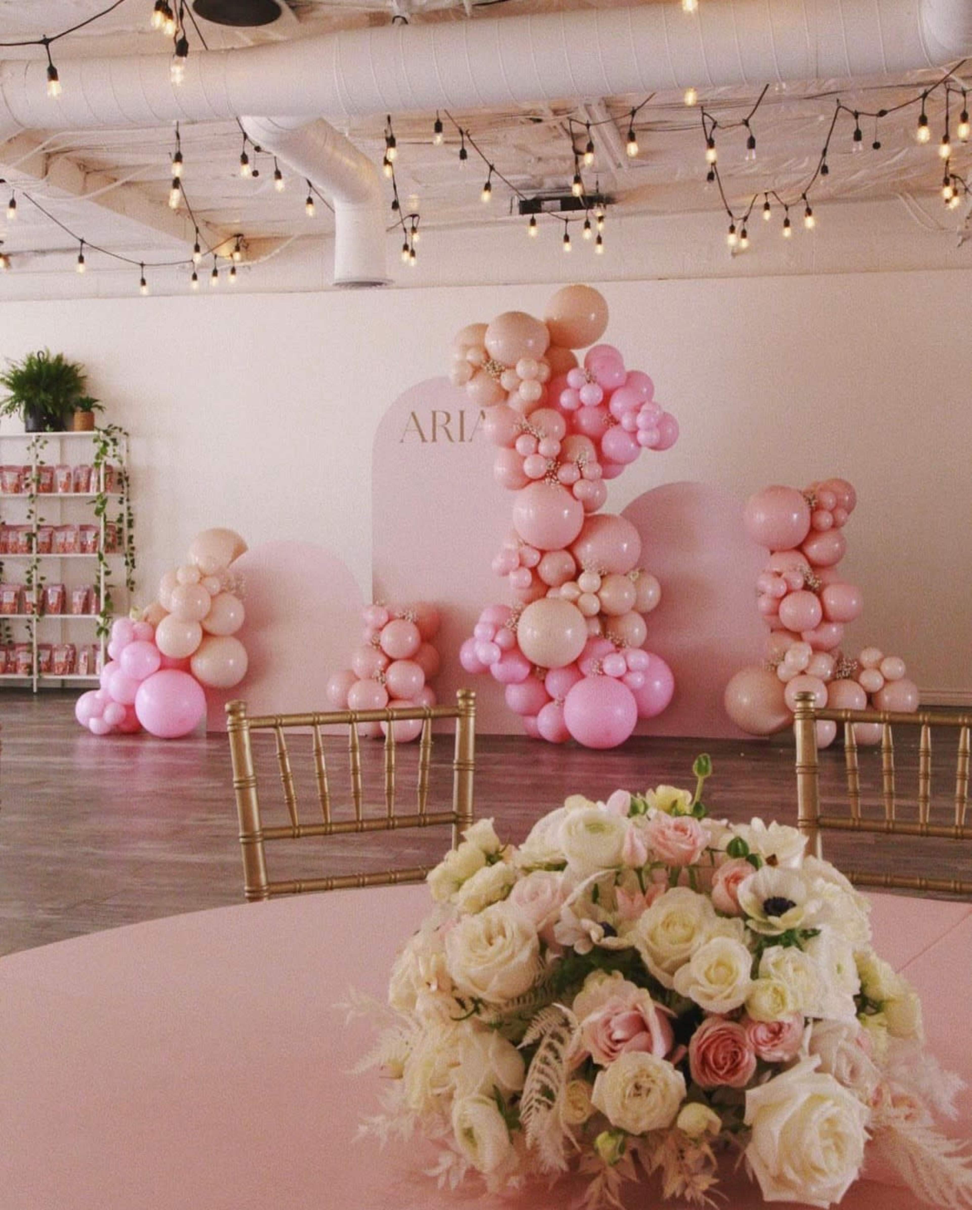 A decorated event space with a backdrop of pink and cream balloons and a floral arrangement on a pink table.