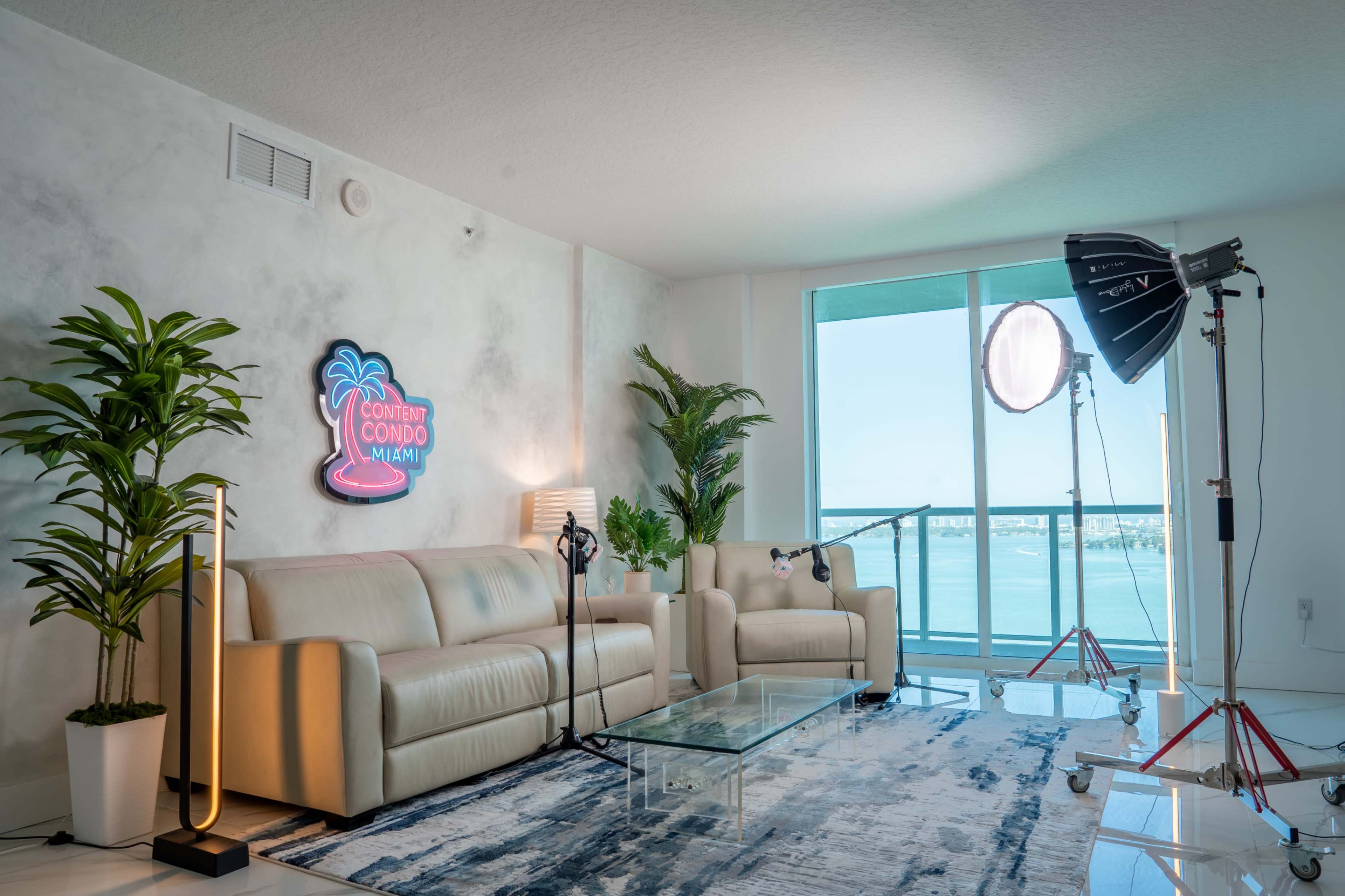 The image shows a modern living room with a glass coffee table, beige couches, indoor plants, and a backdrop of a large window overlooking a body of water.