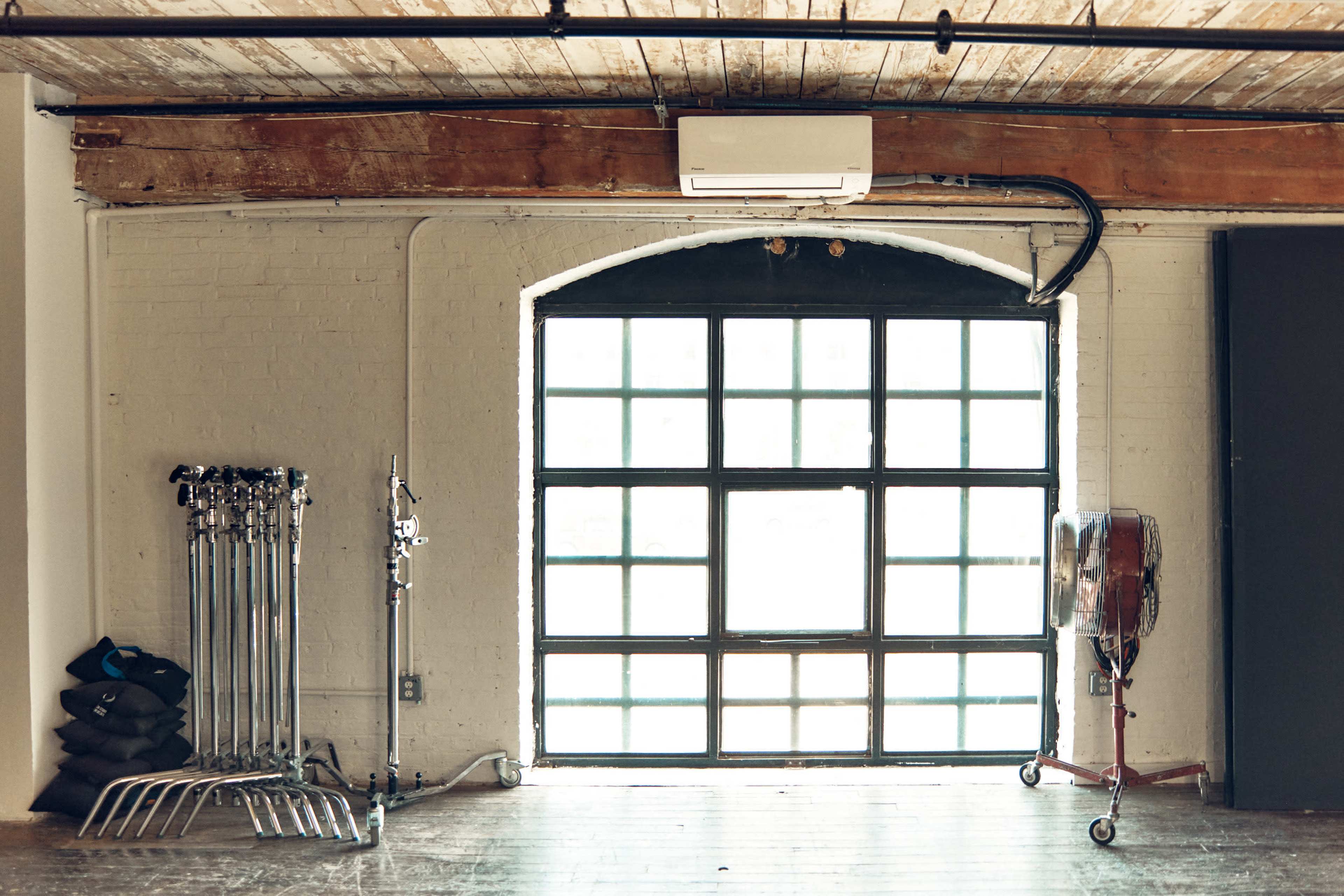 The image shows a spacious room with large windows, a wooden ceiling, and equipment stands lined against one wall.