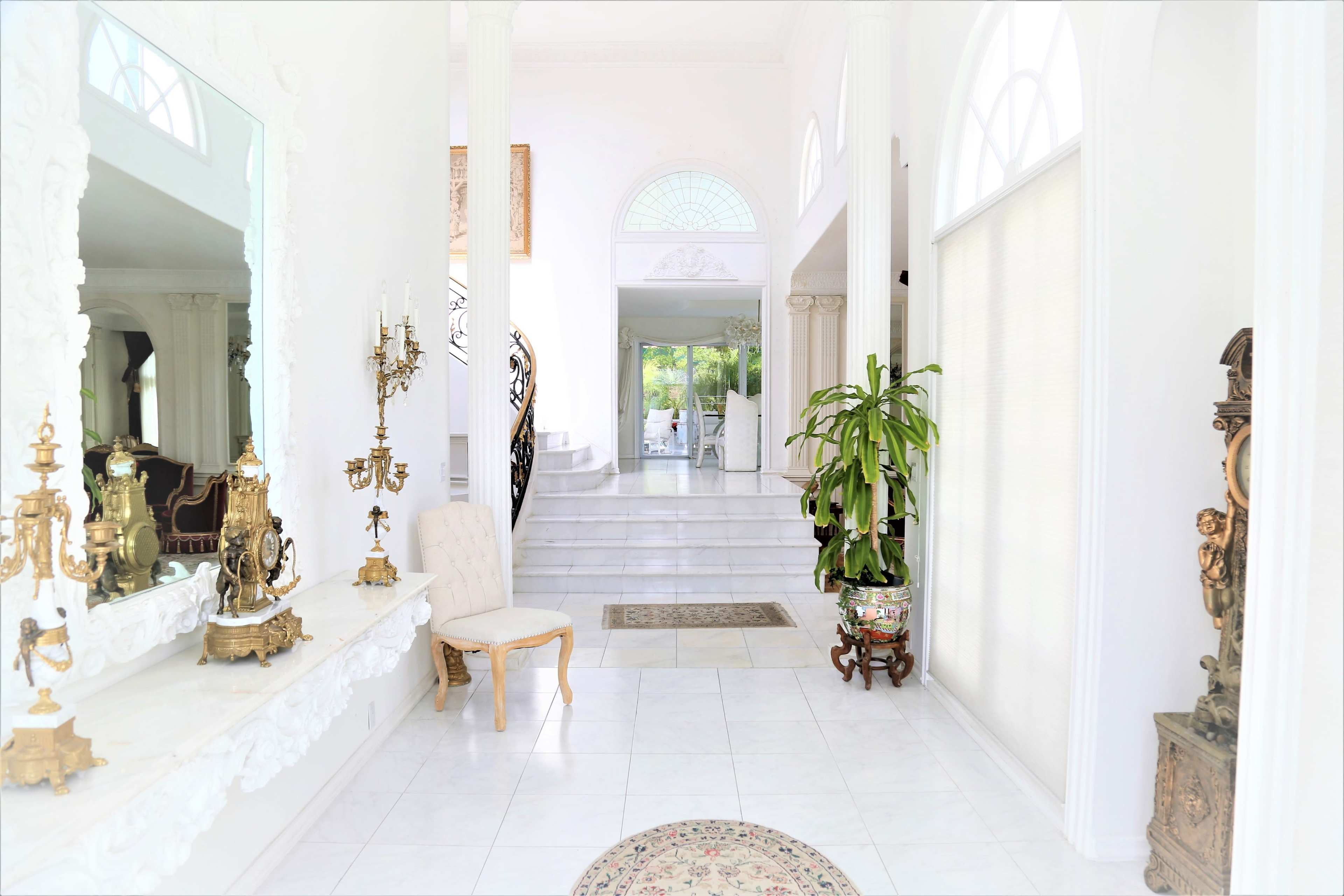 A spacious interior hallway features a large mirror, decorative plants, and ornate furnishings, leading to a staircase and an adjoining room.