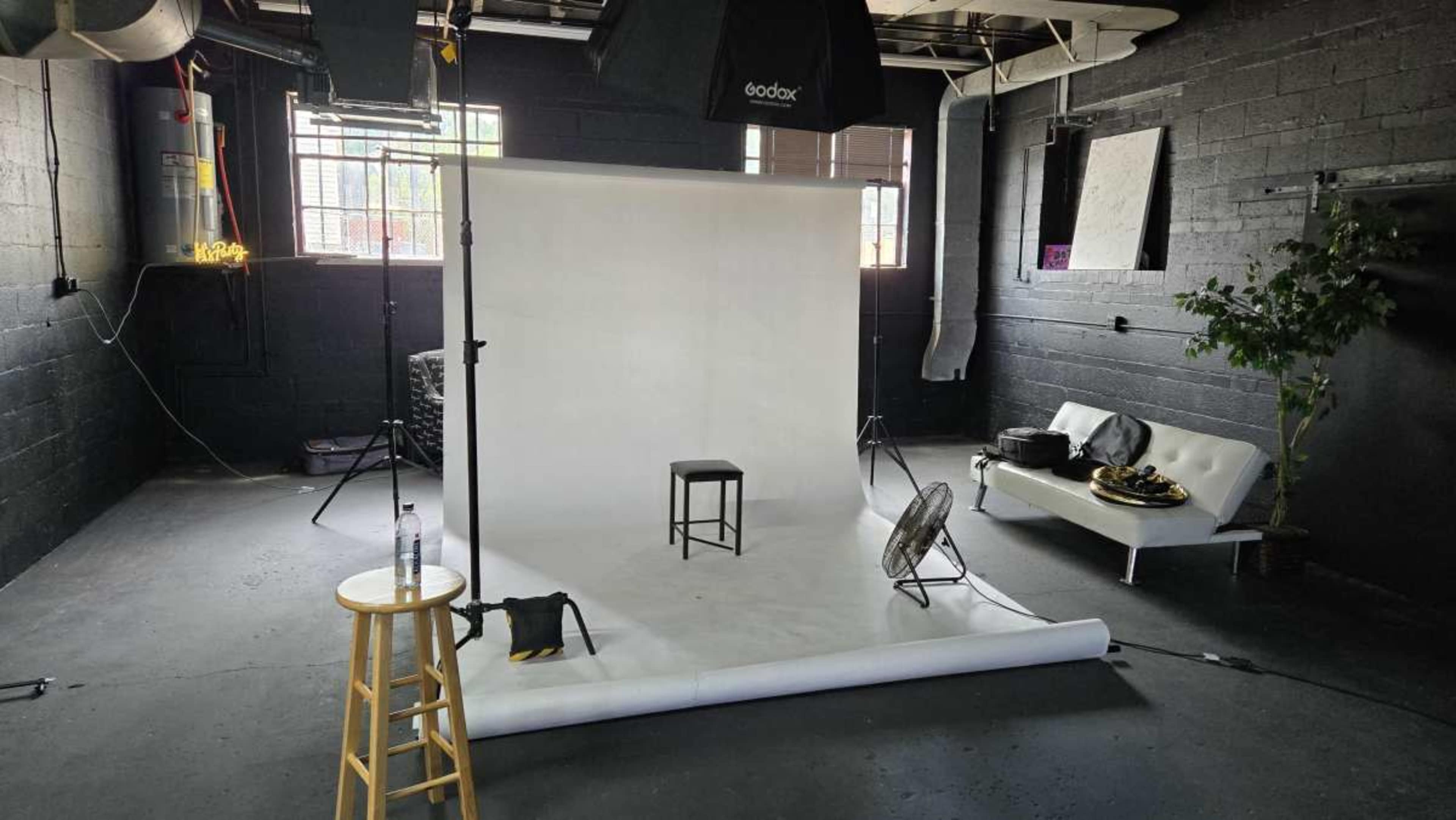 The image shows a photography studio with a white backdrop, a stool in the center, and a couch against the wall, along with lighting equipment and a fan.