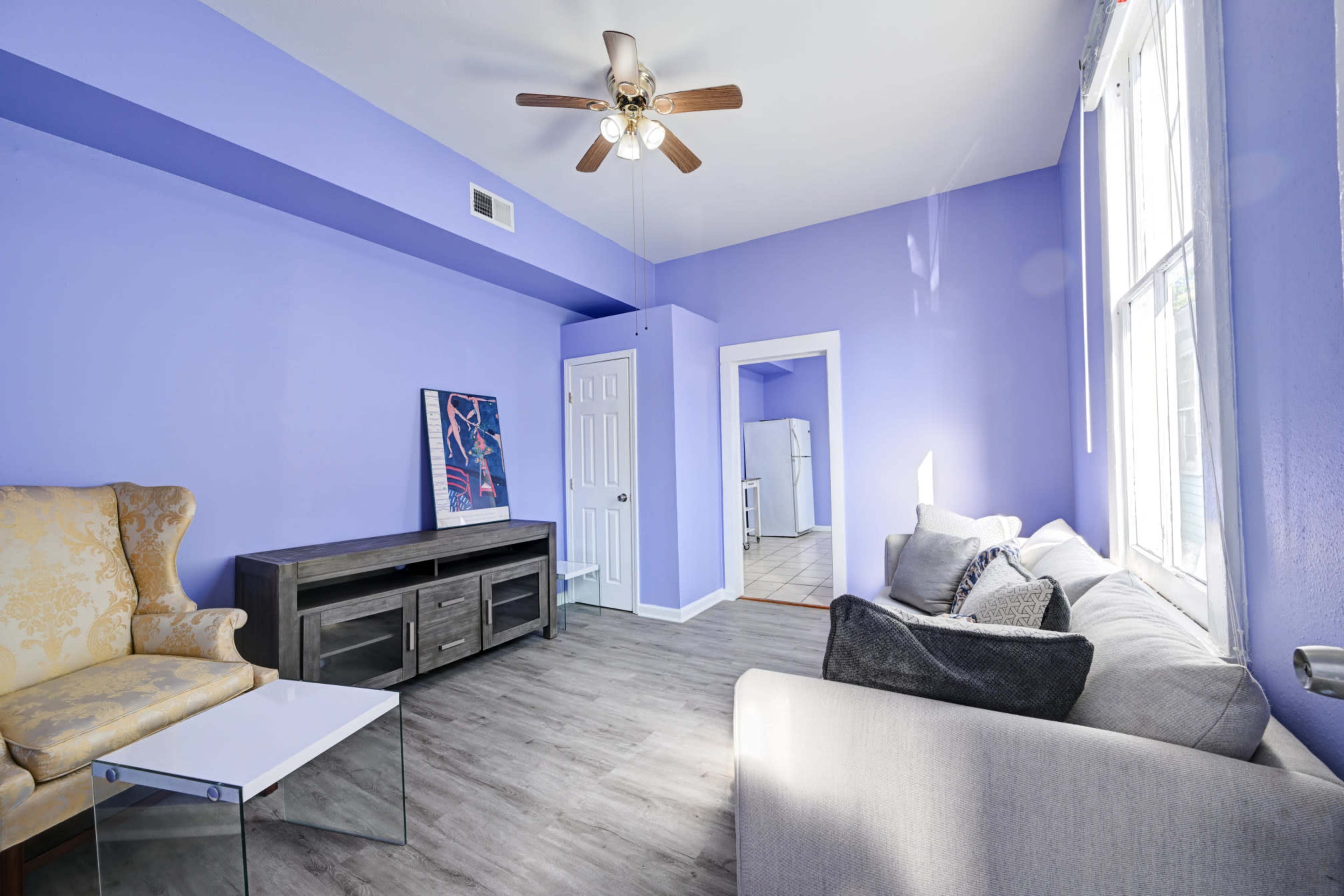 A living room with purple walls, a ceiling fan, and a mix of light furniture including a yellow armchair and a gray sofa.