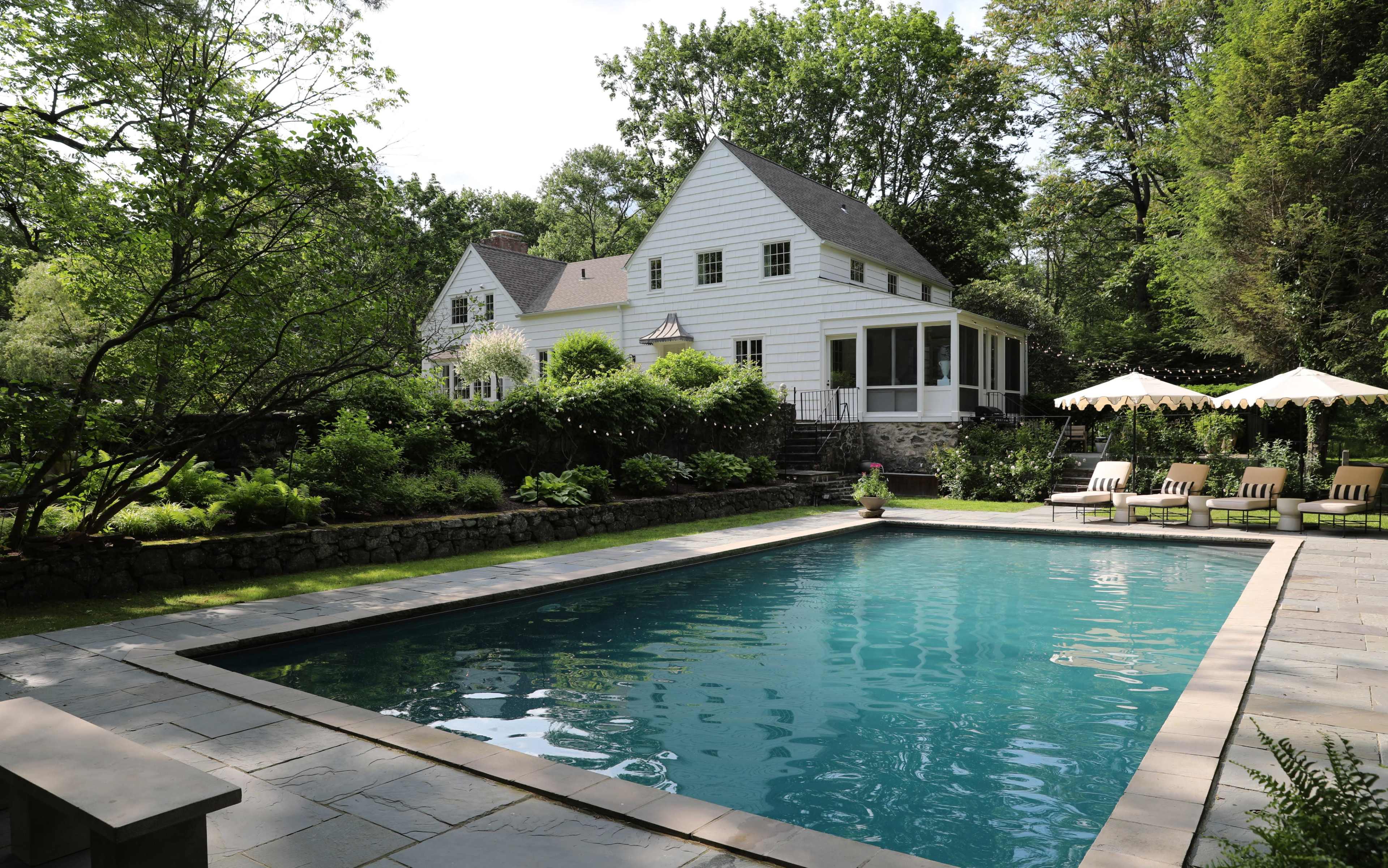 A backyard features a blue swimming pool surrounded by a stone patio, with lounge chairs, lush greenery, and a white house in the background.