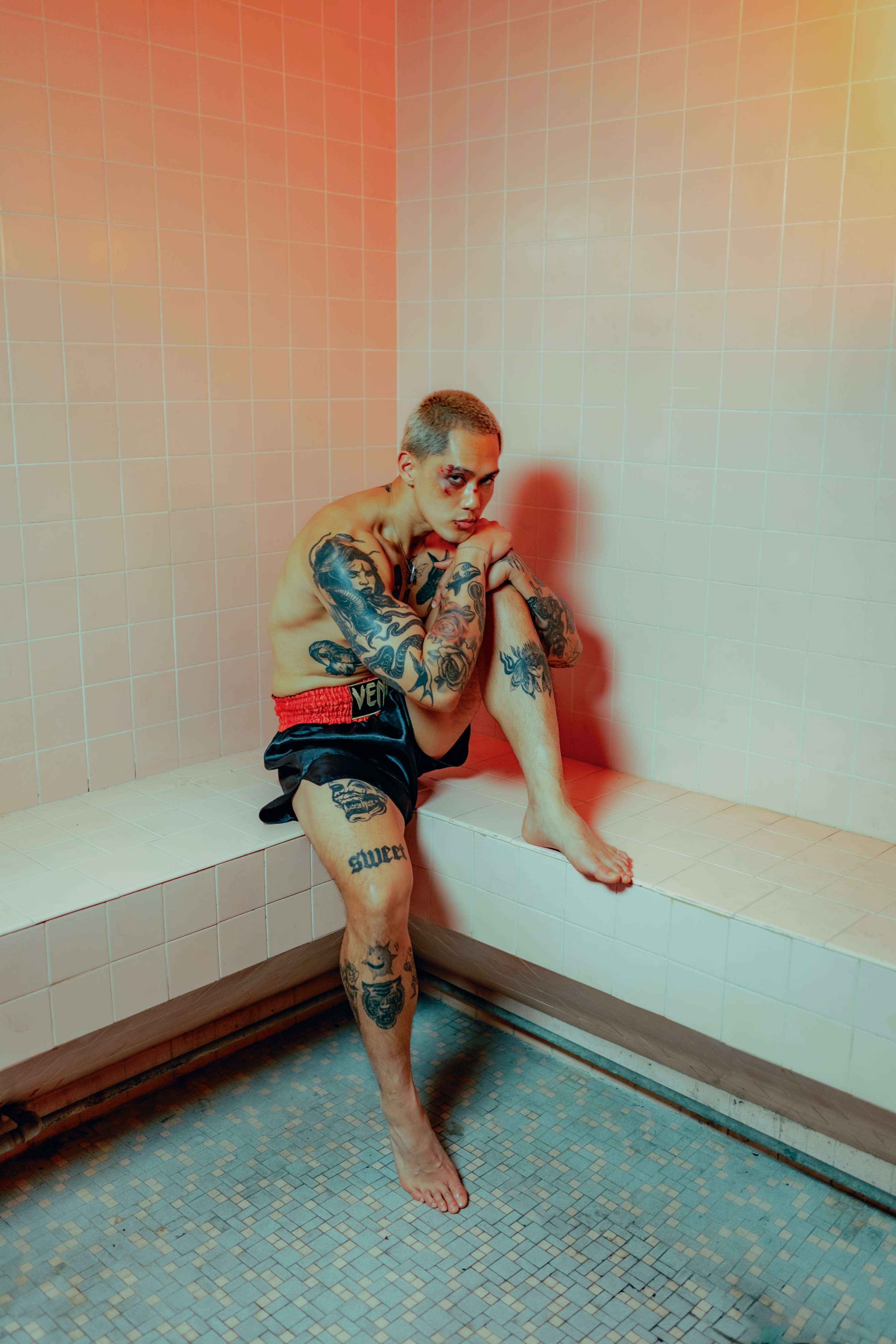 A tattooed individual sits on a tiled bench in a softly lit bathroom space.