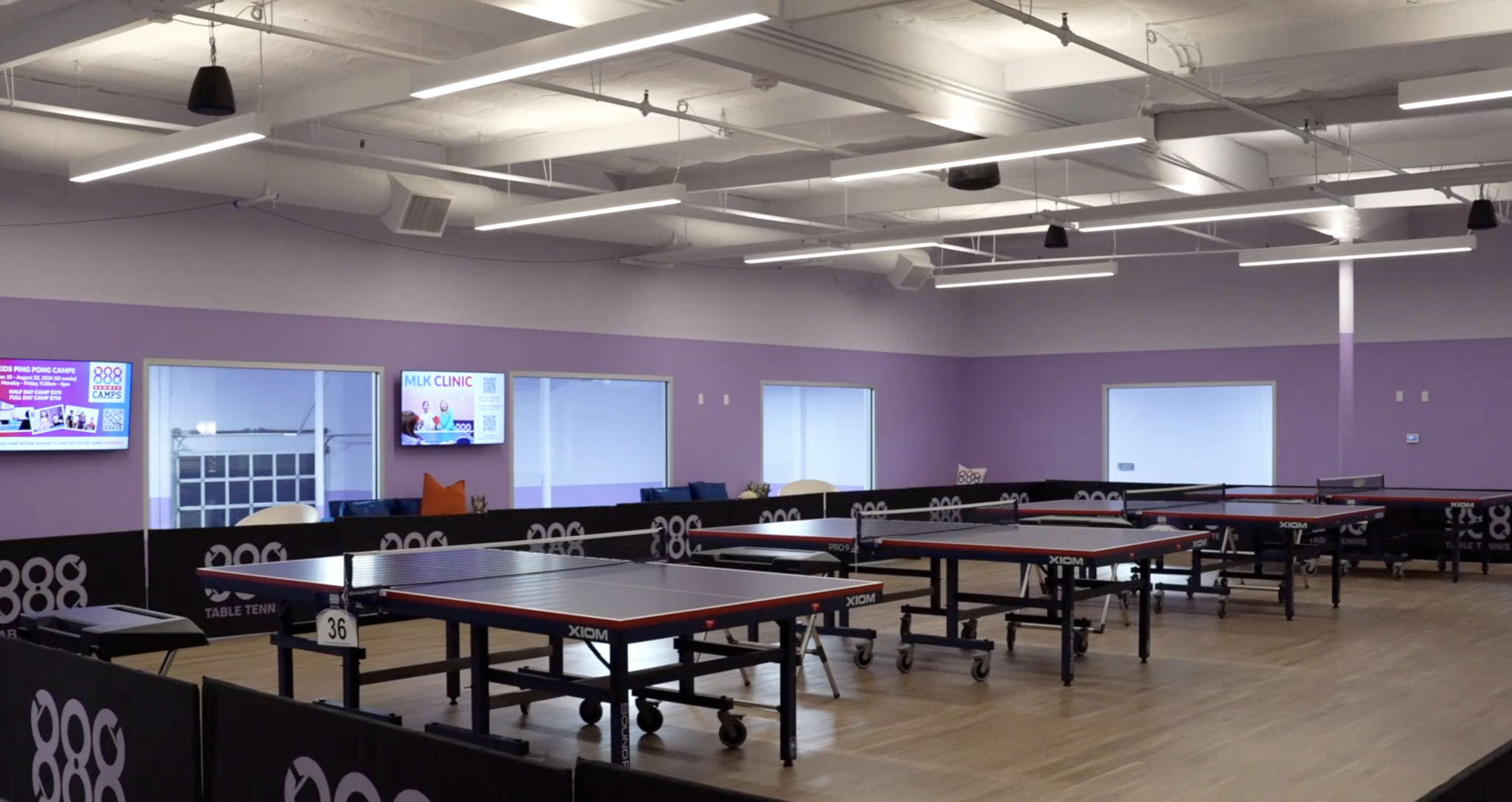 The image shows an indoor table tennis facility with several tables arranged in a spacious room featuring purple walls and bright overhead lighting.